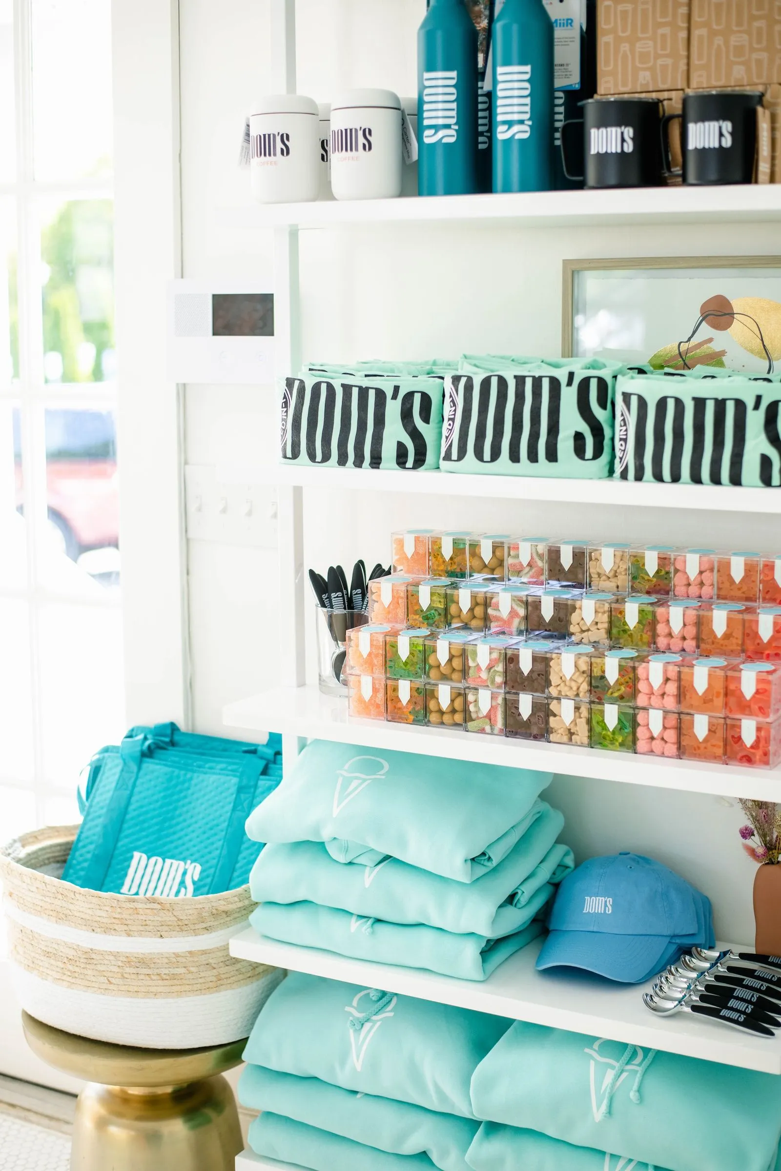 Organized retail shelving unit displaying Dom's branded merchandise, including packaged goods, drink containers, apparel, and accessories, showcasing the brand's product range and visual identity.