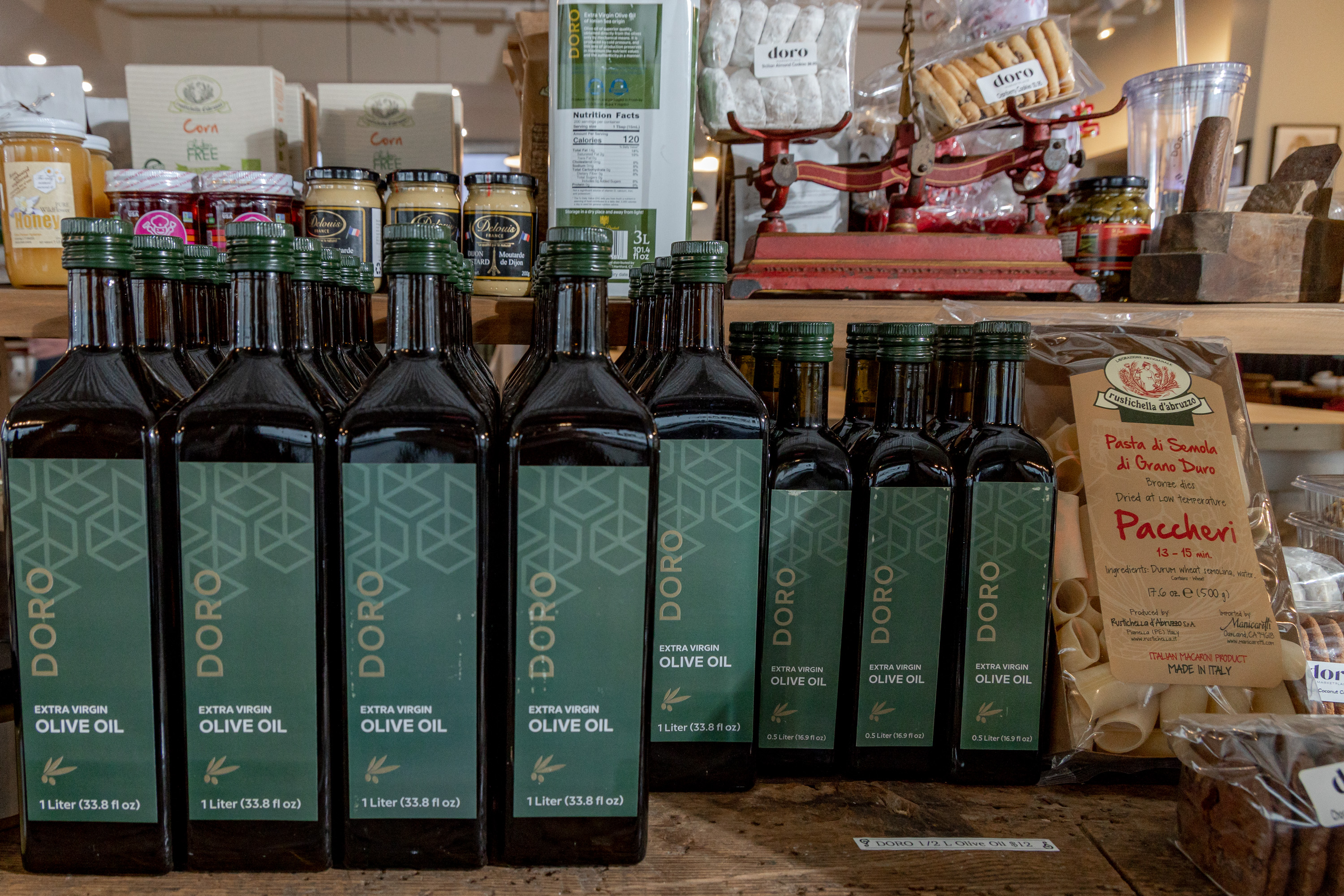 A photo showcasing a display of Doro Extra Virgin Olive Oil bottles on a shelf at the Doro Marketplace. The bottles are neatly arranged alongside other gourmet food items, creating a visually appealing and inviting display.
