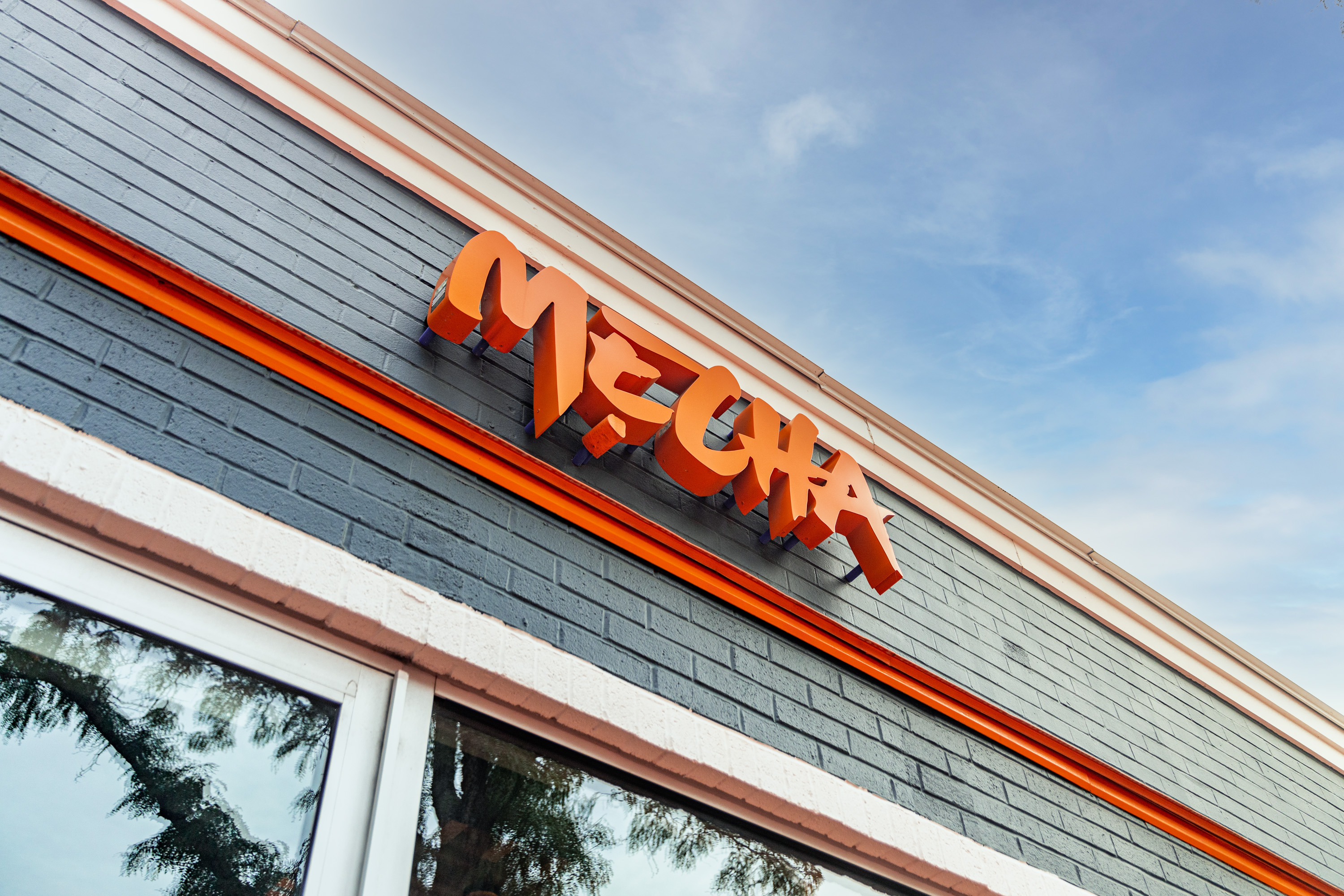 Angled view of the Mecha restaurant exterior, highlighting the dimensional, orange logo signage mounted on a gray brick facade with contrasting orange trim.