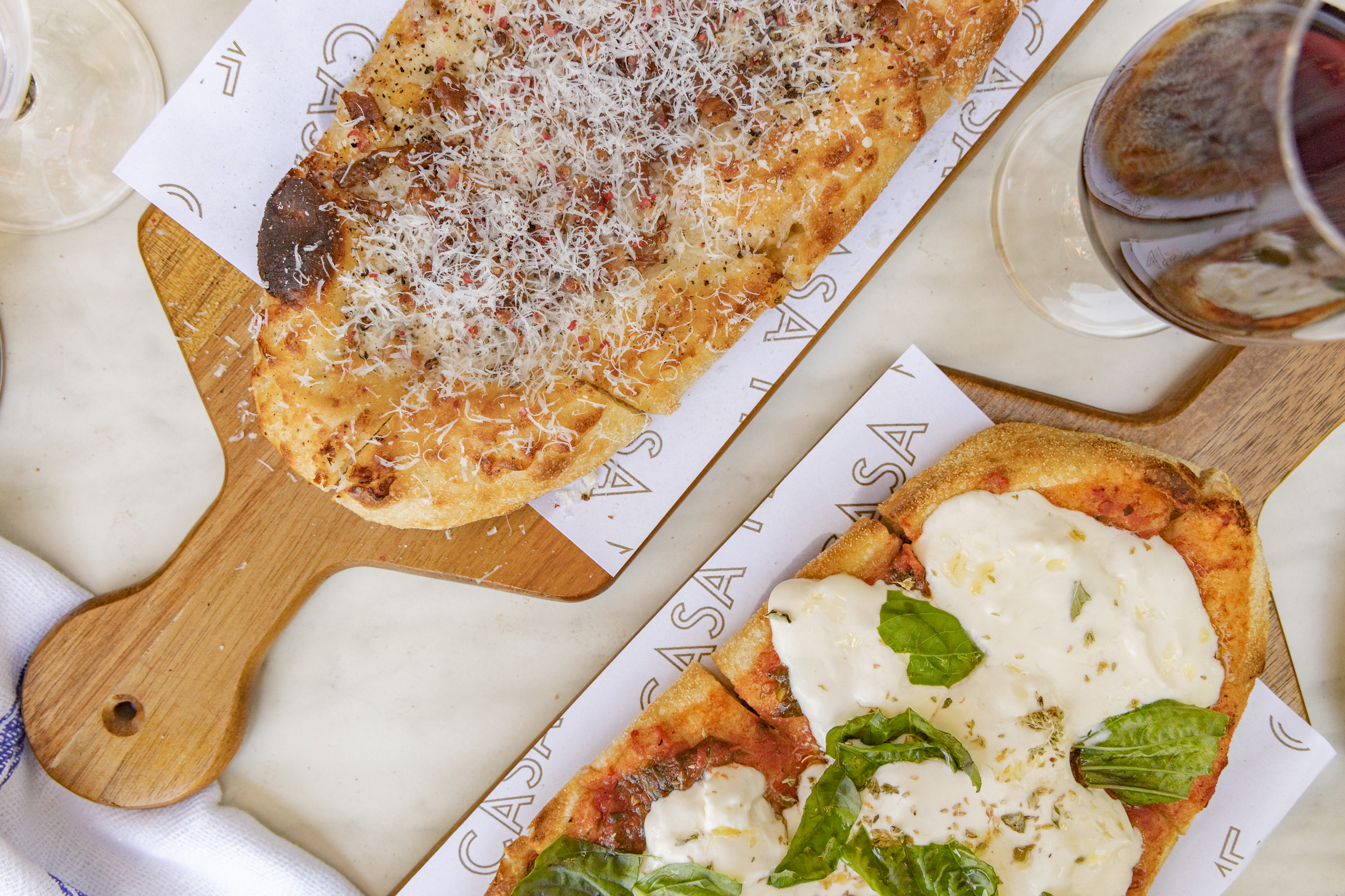 Image of two artisanal pizzas highlighting their varied toppings, textures, and the Casa Me branding through printed liners, emphasizing a rustic and appetizing presentation.