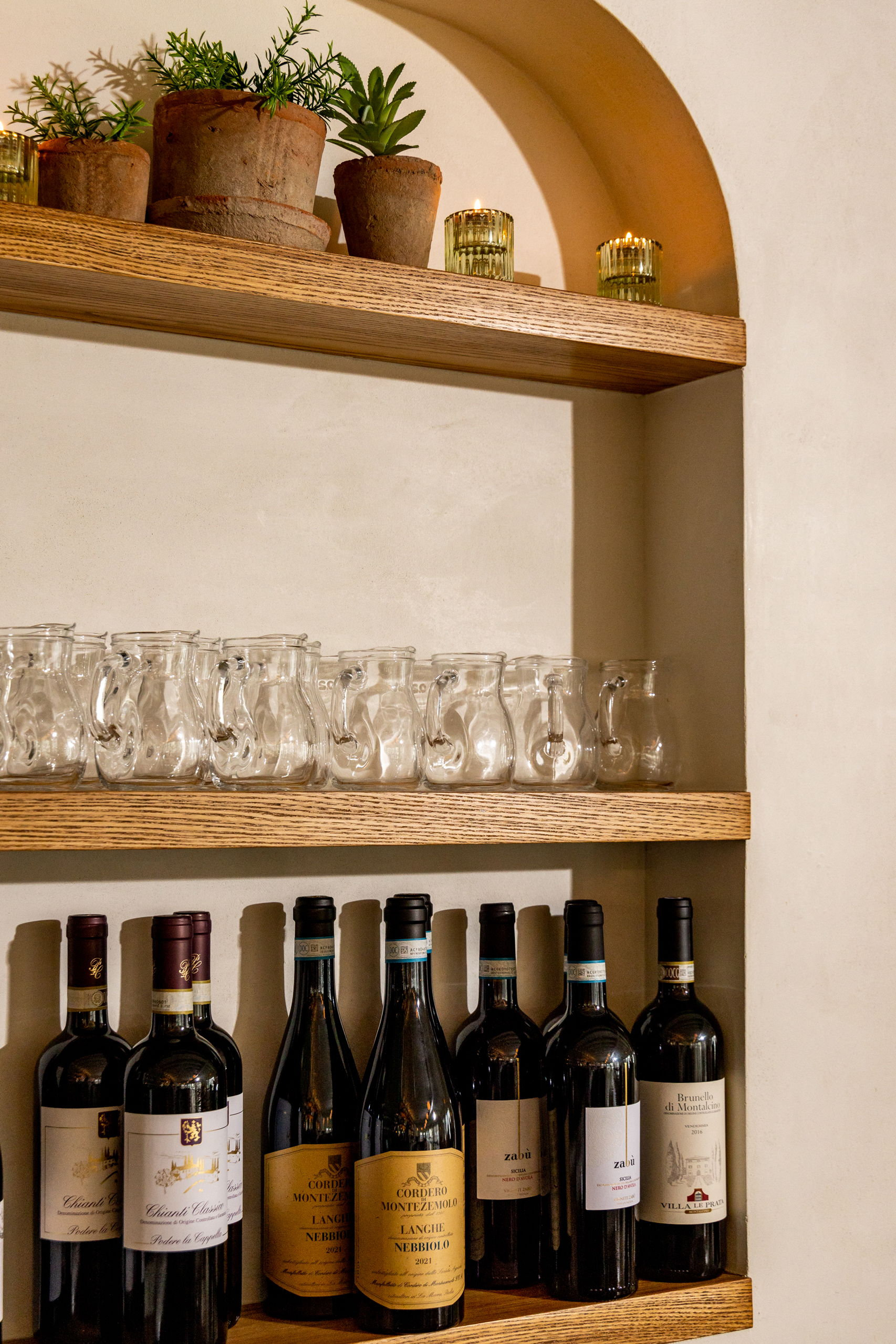 Image of neatly arranged wine bottles on a lower shelf and glassware on an upper shelf, showcasing a rustic display with warm lighting and a focus on product presentation.