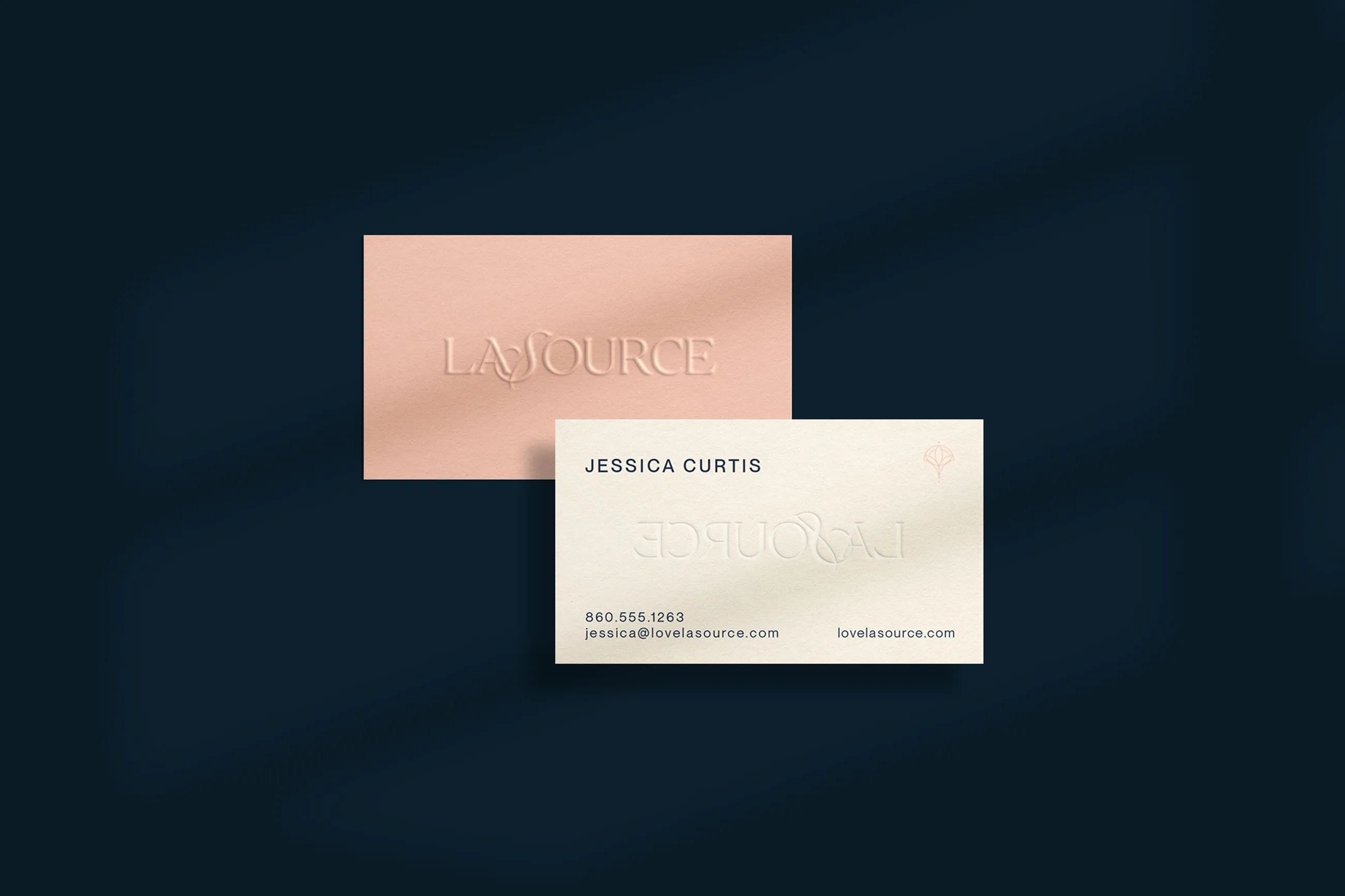 Two business cards for LASOURCE, one peach-colored card lying flat with an embossed logo, and a white card angled on top featuring contact information and a blind embossed logo, all against a dark blue background.