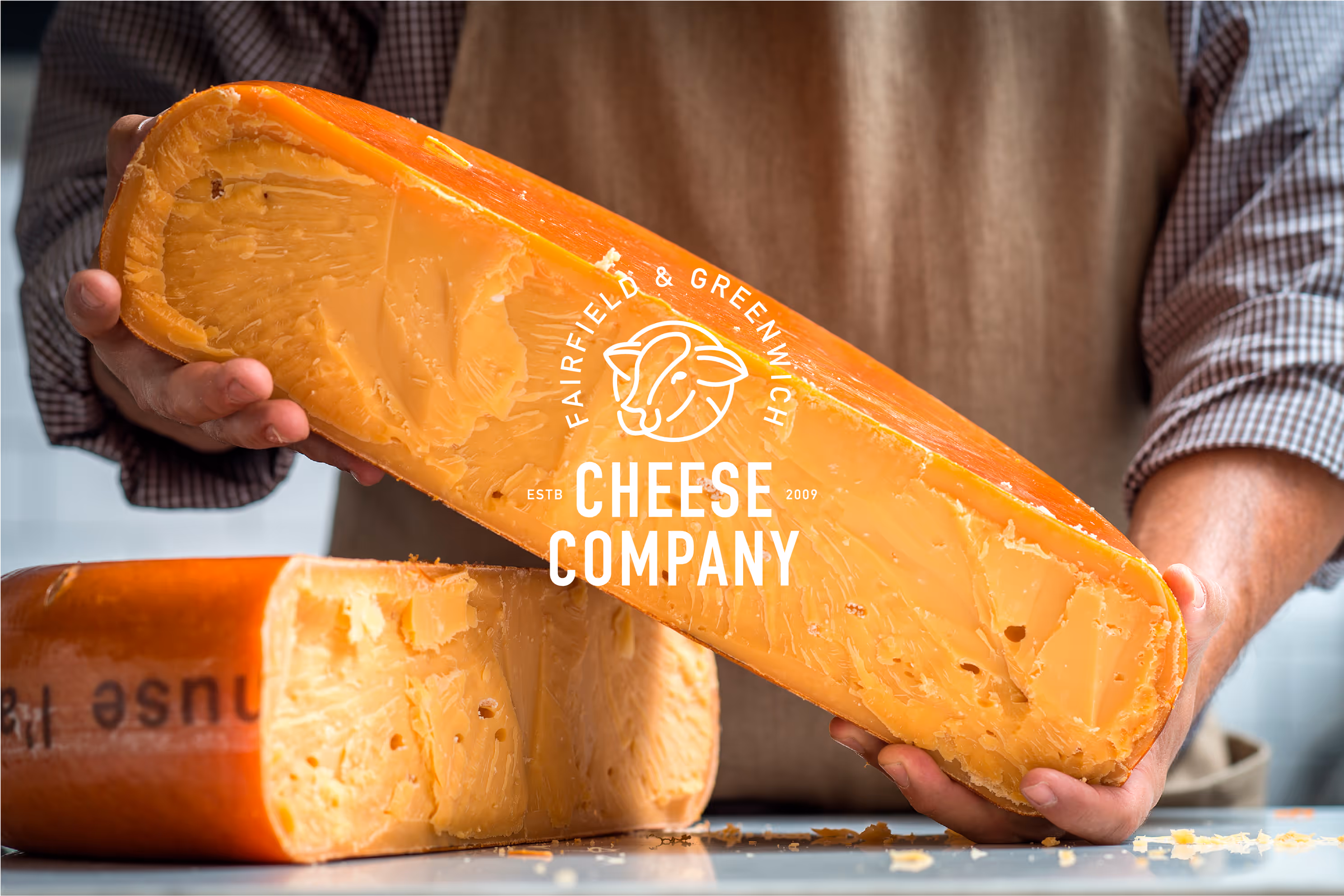 Image of two blocks of orange cheese, one held by a person wearing an apron, showcasing the Fairfield Cheese Company logo and branding.