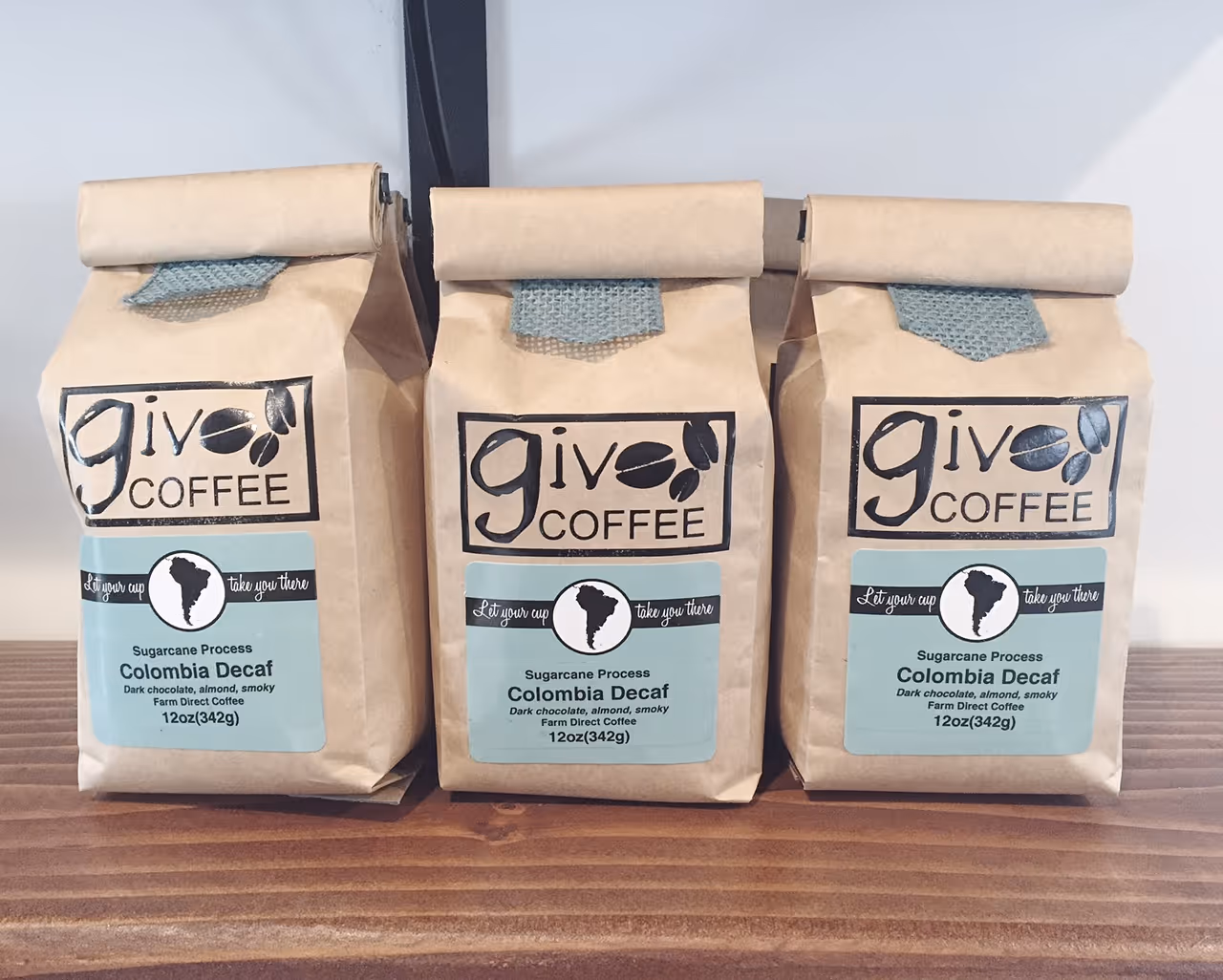 Three kraft bags of Giv Coffee's Colombia Decaf, featuring a minimalist design with black logo, teal label, and natural fabric tab closure, sit on a wooden shelf.