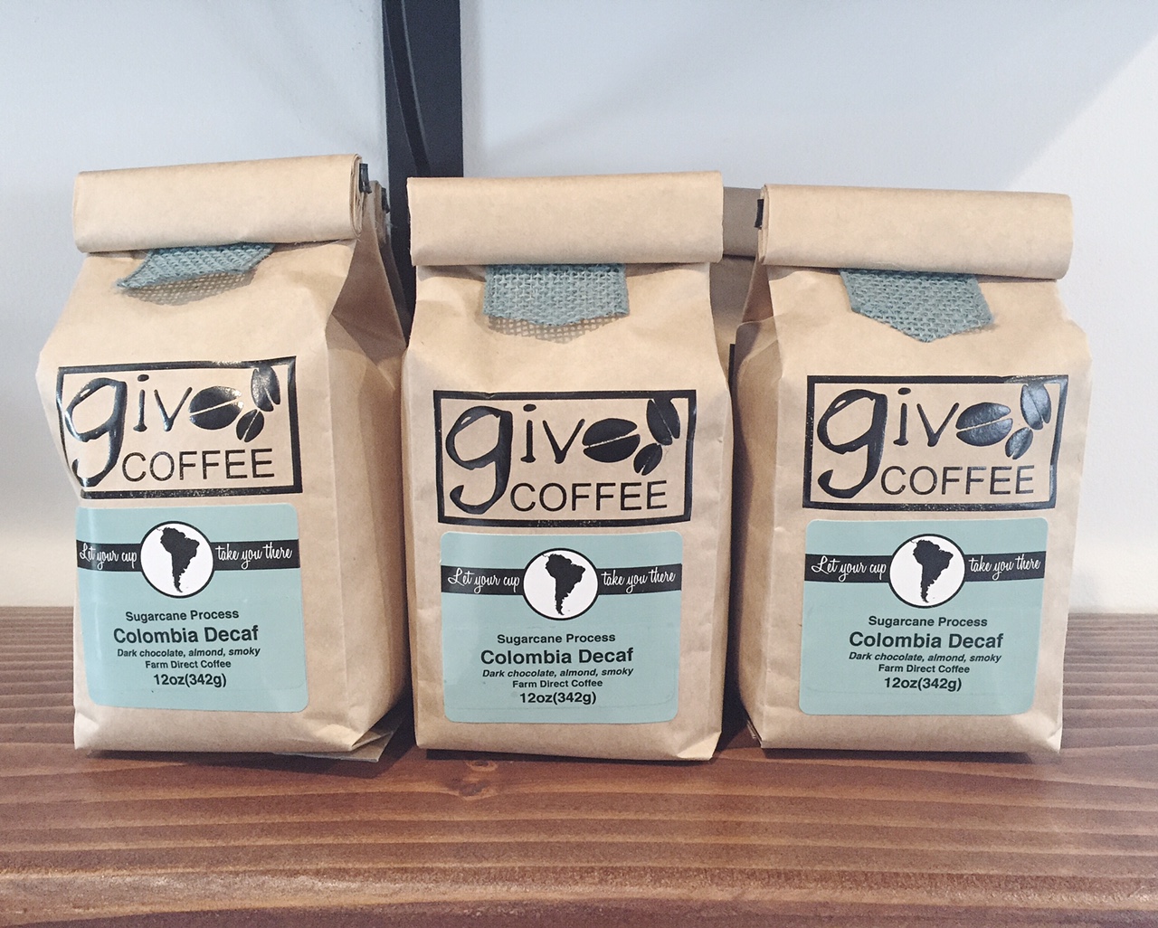 Three kraft bags of Giv Coffee's Colombia Decaf, featuring a minimalist design with black logo, teal label, and natural fabric tab closure, sit on a wooden shelf.