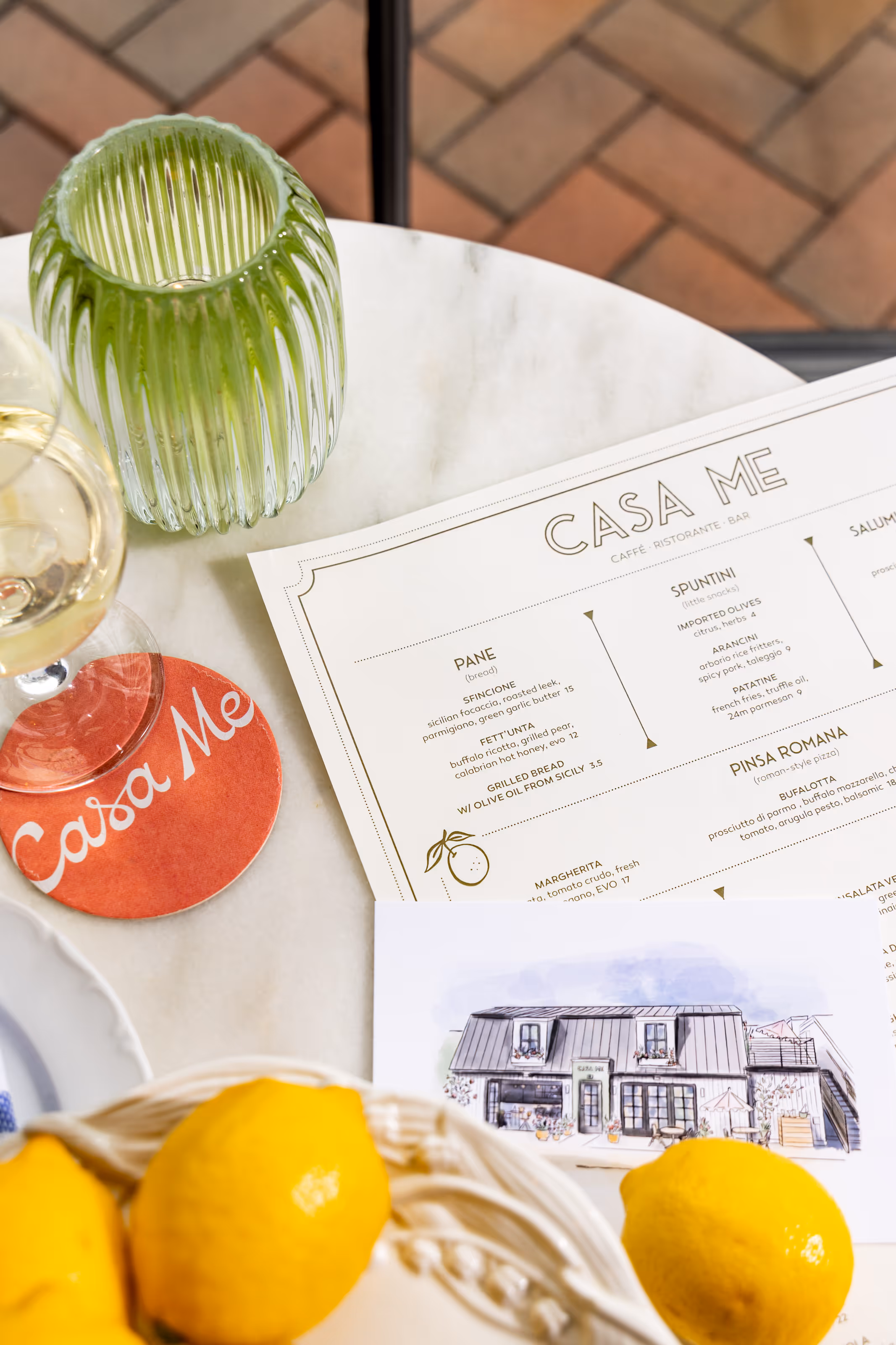 Flat lay of a Casa Me menu, coaster, illustrated card, lemons, and a glass container, showcasing the restaurant's branding and table presentation on a marble surface.