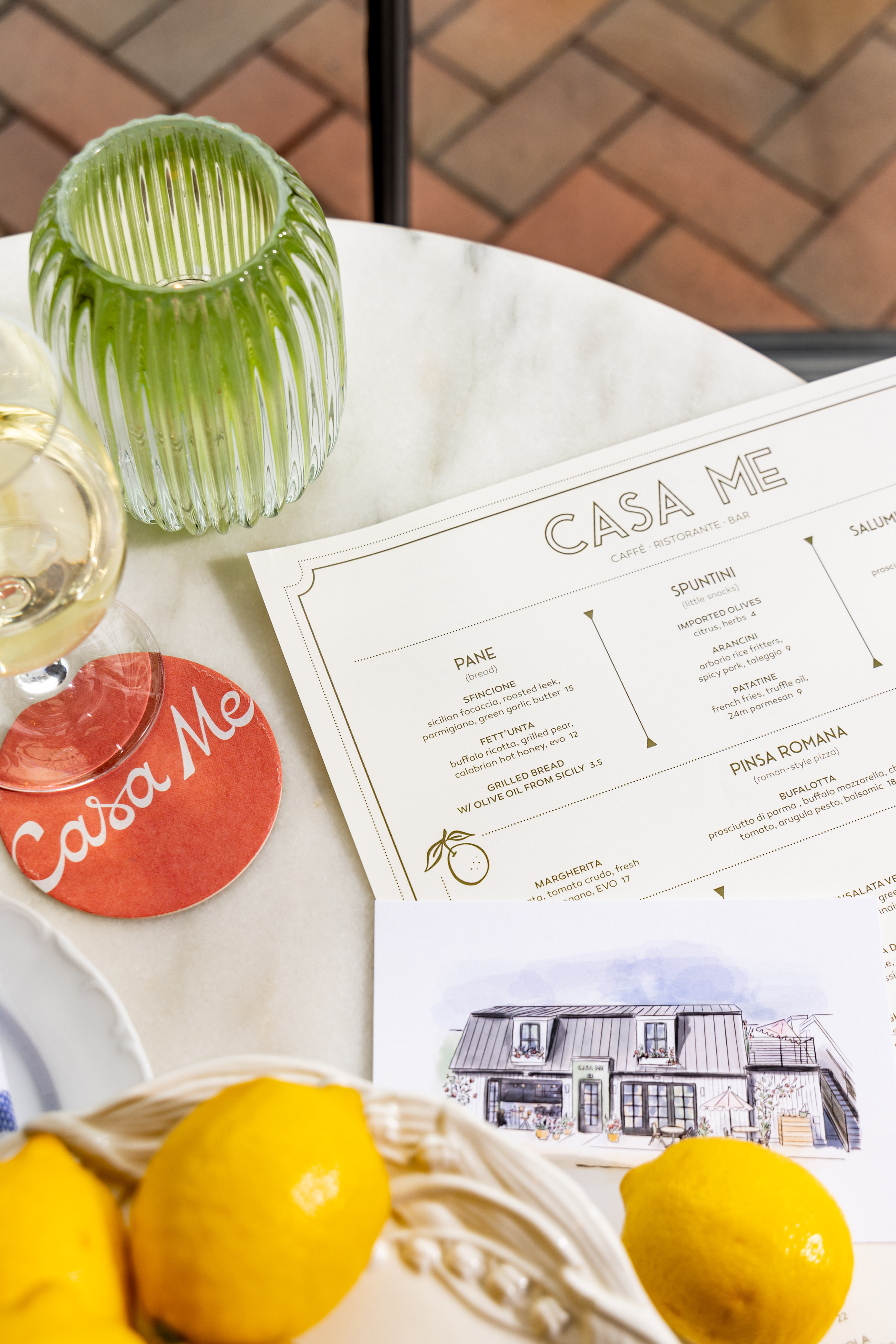 Flat lay of a Casa Me menu, coaster, illustrated card, lemons, and a glass container, showcasing the restaurant's branding and table presentation on a marble surface.