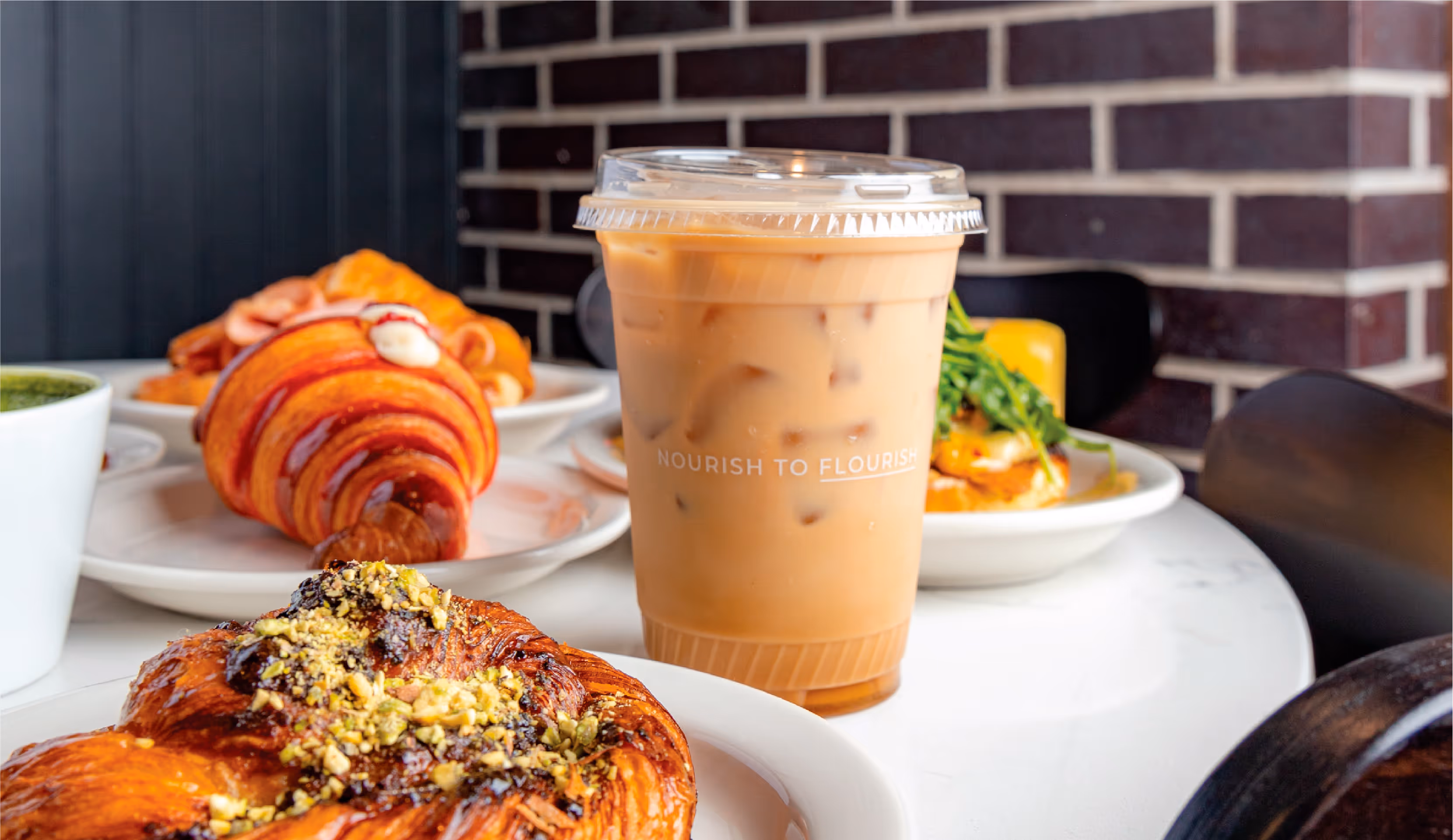 A close-up photo of an iced coffee drink in a clear plastic cup, surrounded by plates of pastries on a cafe table. The "Nourish to Flourish" text on the cup adds a touch of branding to the image.