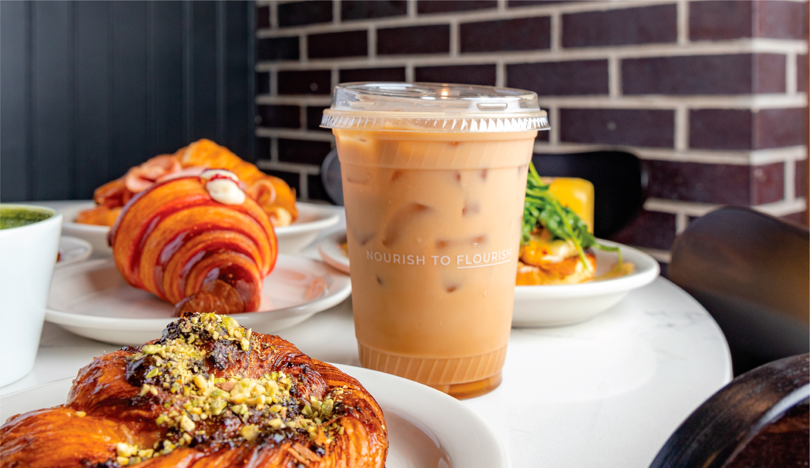 A close-up photo of an iced coffee drink in a clear plastic cup, surrounded by plates of pastries on a cafe table. The "Nourish to Flourish" text on the cup adds a touch of branding to the image.
