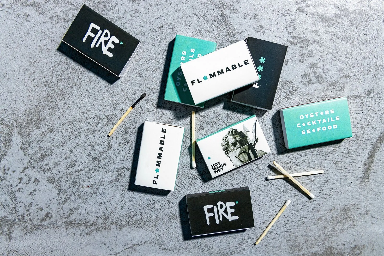 Collection of RAW matchboxes, highlighting the brand's eclectic design approach. The matchboxes feature contrasting color palettes, bold typography, and playful graphics, ranging from fire and safety themes to a seafood-focused design with an oyster graphic.