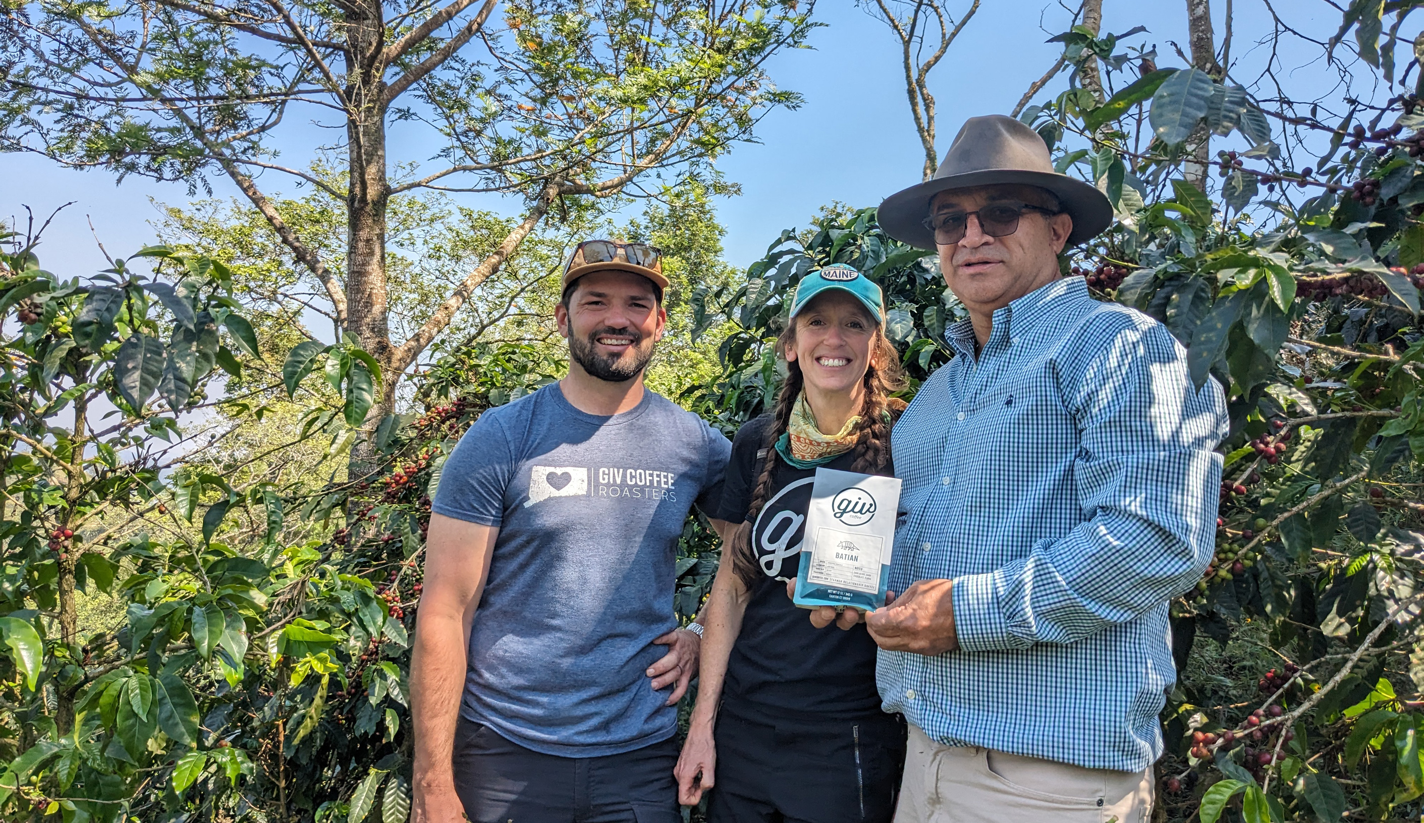 Three people stand amongst coffee plants, one holding a bag of Giv Coffee, highlighting the origin of the coffee.