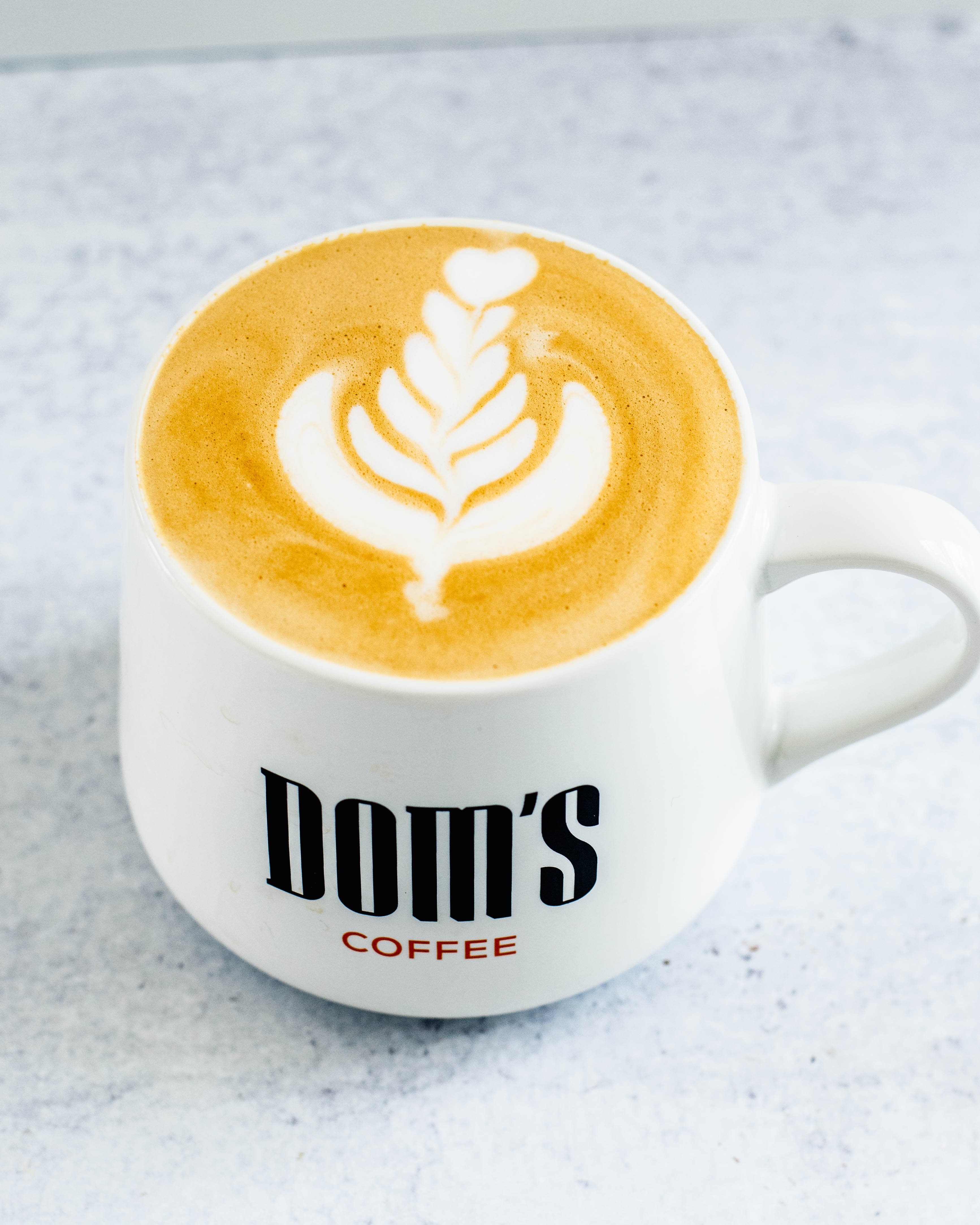 Image showcasing latte art in a white cup, highlighting the intricate design created in the coffee foam and the branding of "Dom's Coffee" through typography on the cup.