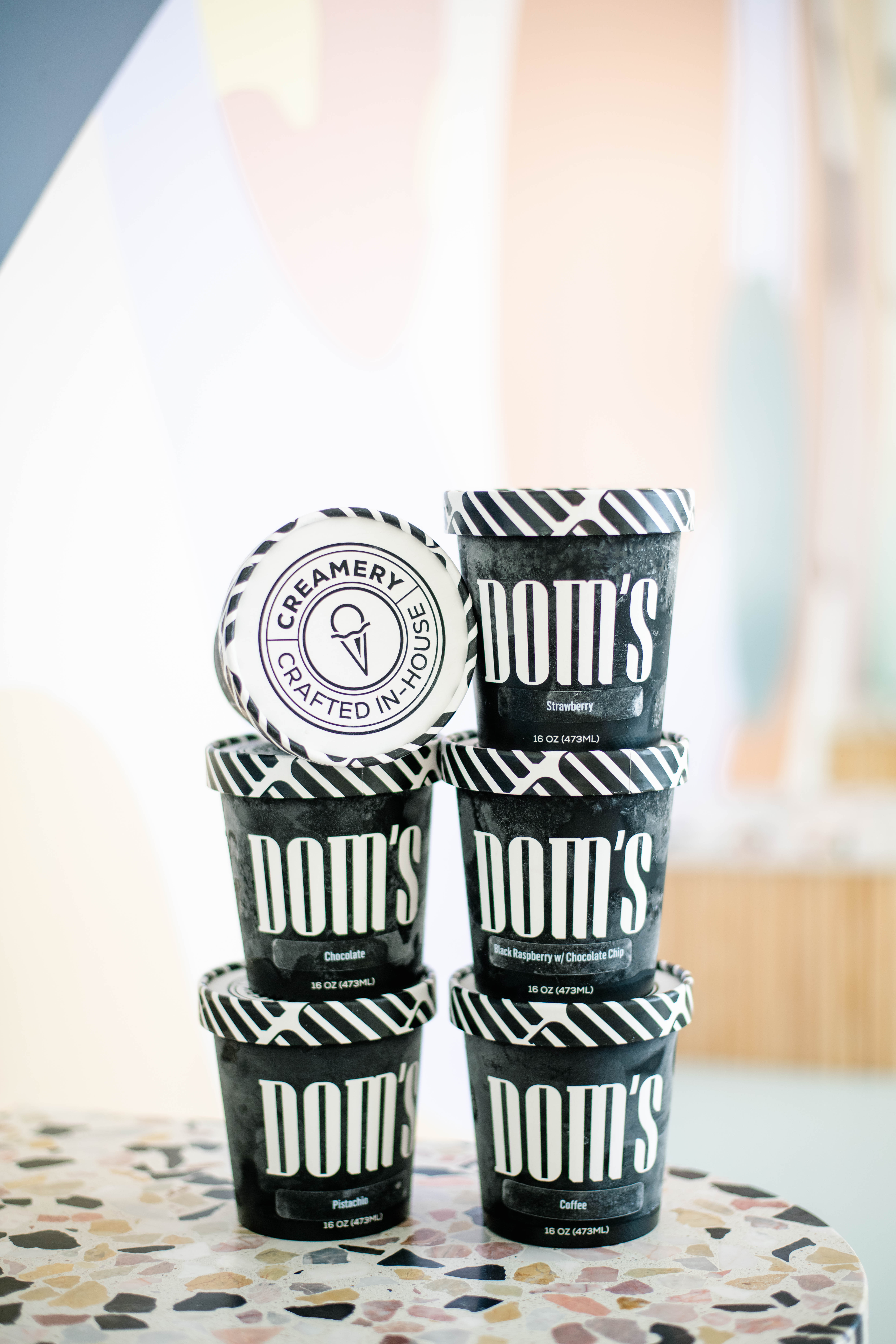 Stack of black ice cream containers with the "Dom's" logo and flavor labels, including a lid with the "Creamery Crafted In-House" branding, displayed on a speckled surface.