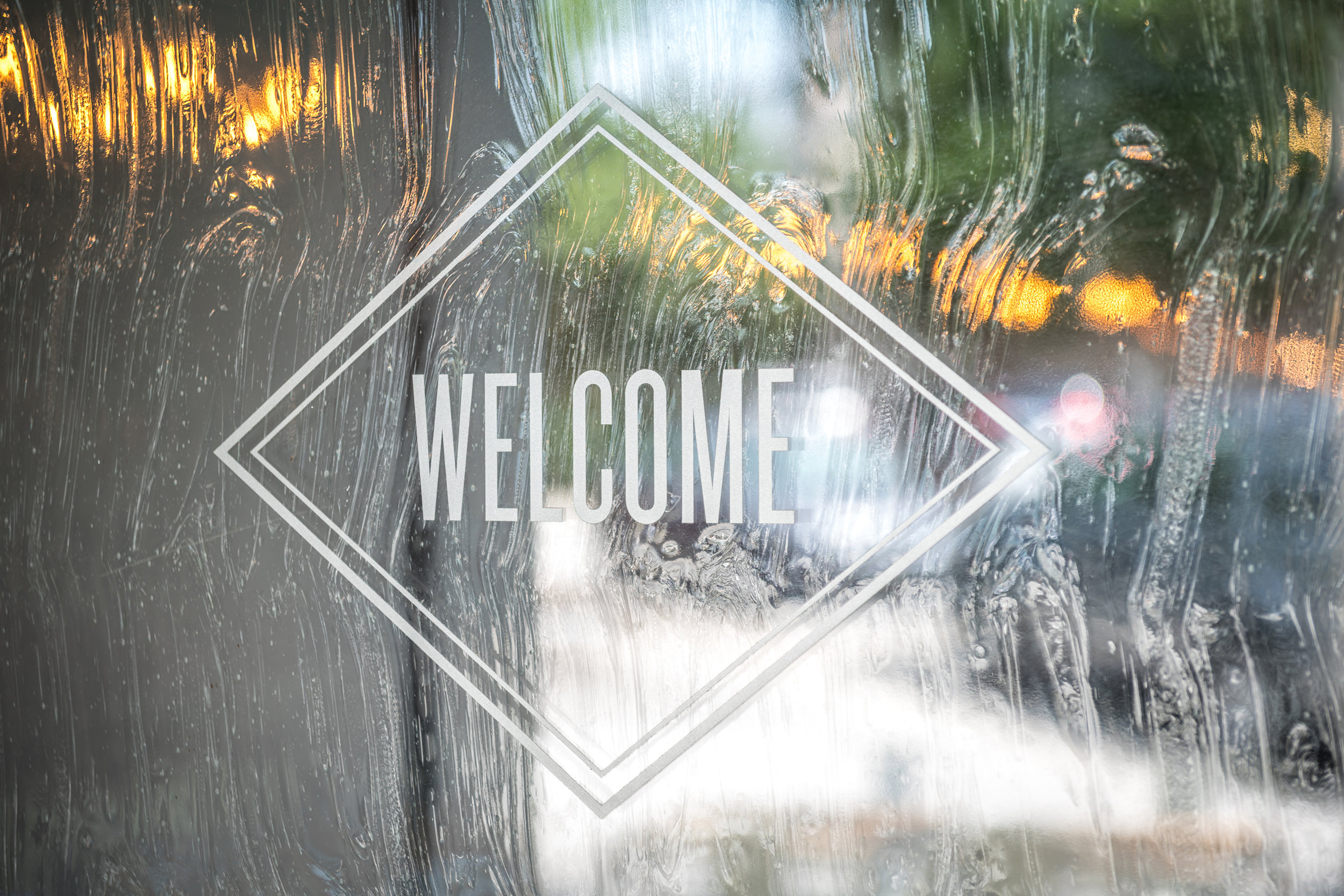 A simple yet effective 'Welcome' sign design, using a geometric diamond shape to frame the word. The rain-streaked glass adds a textured background.