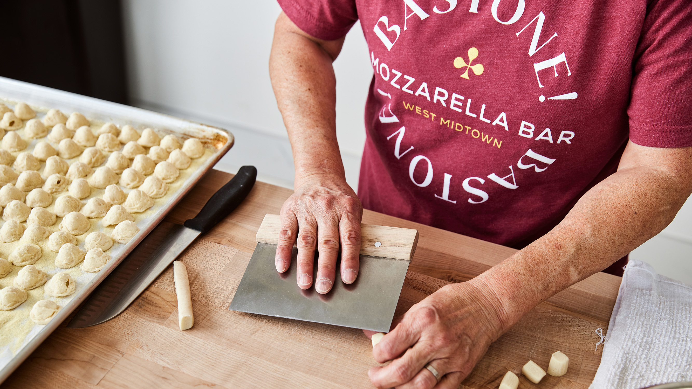 A close-up photo of a chef's hands skillfully shaping and cutting fresh mozzarella cheese on a wooden board. The chef wears a t-shirt emblazoned with the "Bastone! Mozzarella Bar" logo, highlighting the brand's focus on artisanal cheesemaking.
