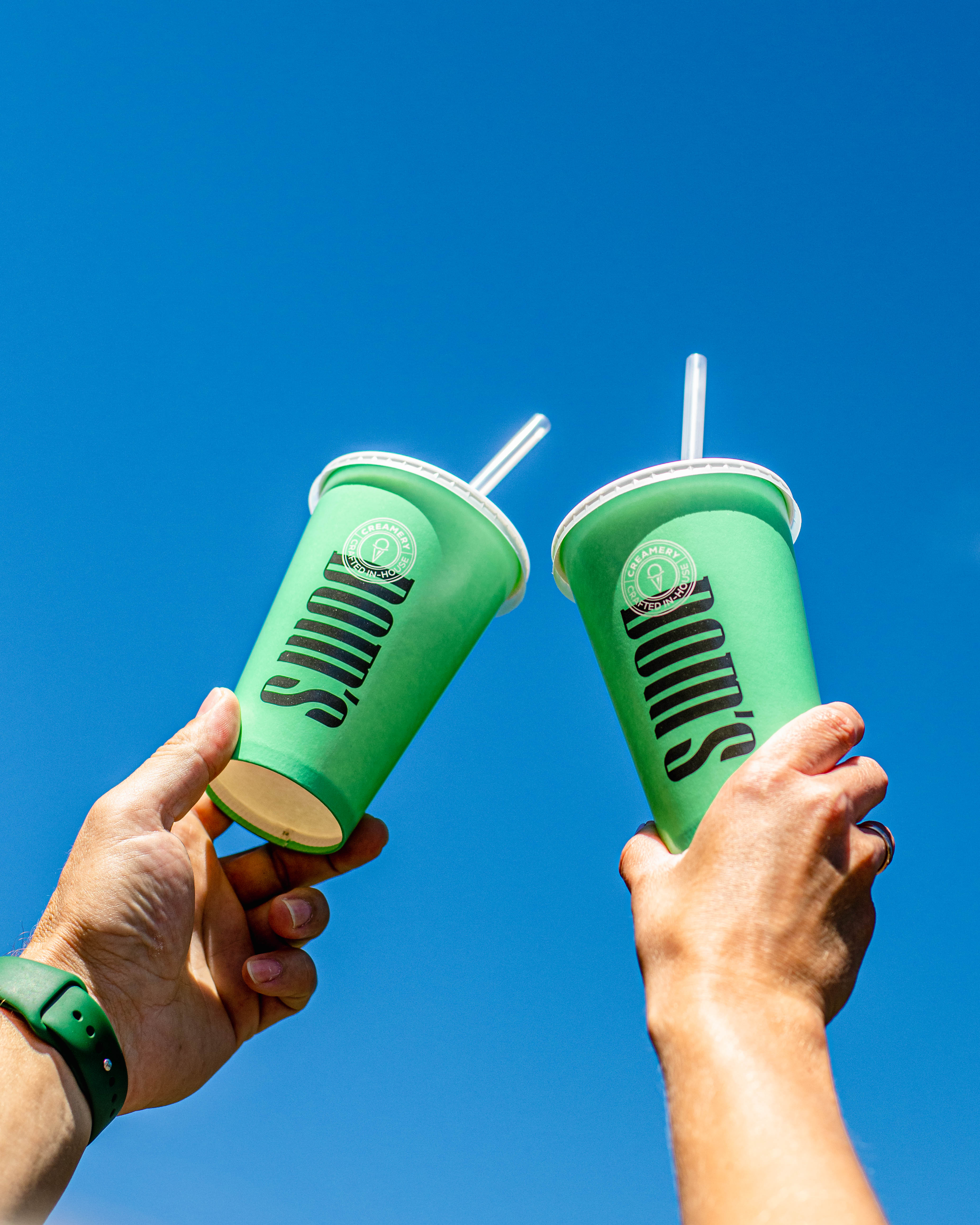 Close-up of two hands holding matching light green Dom's cups with white straws against a bright blue sky, showcasing the cups' design and branding.