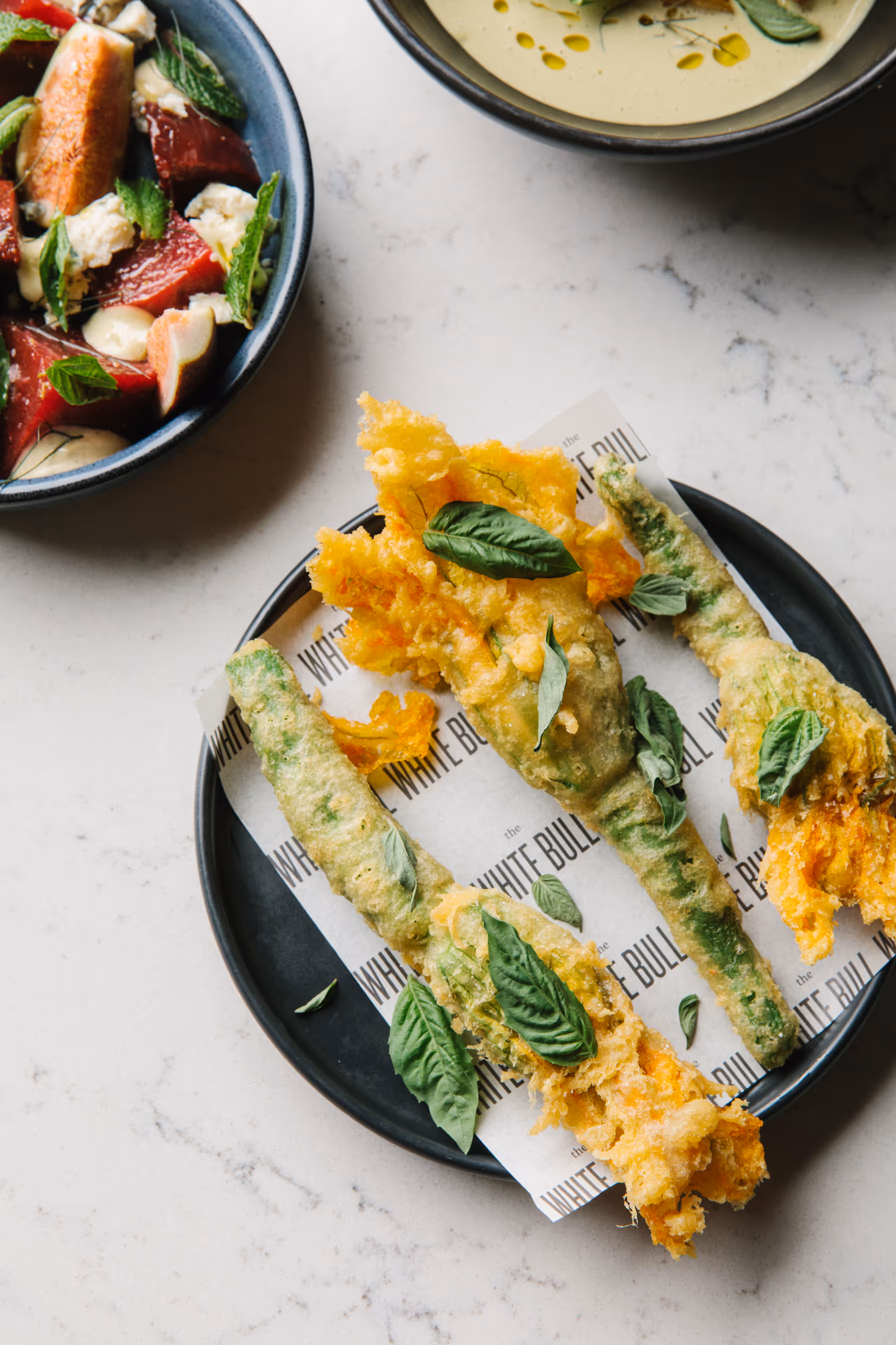 Three fried squash blossoms, garnished with fresh basil, served on a black plate lined with paper printed with 'The White Bull' logo. Other dishes are visible in the background.