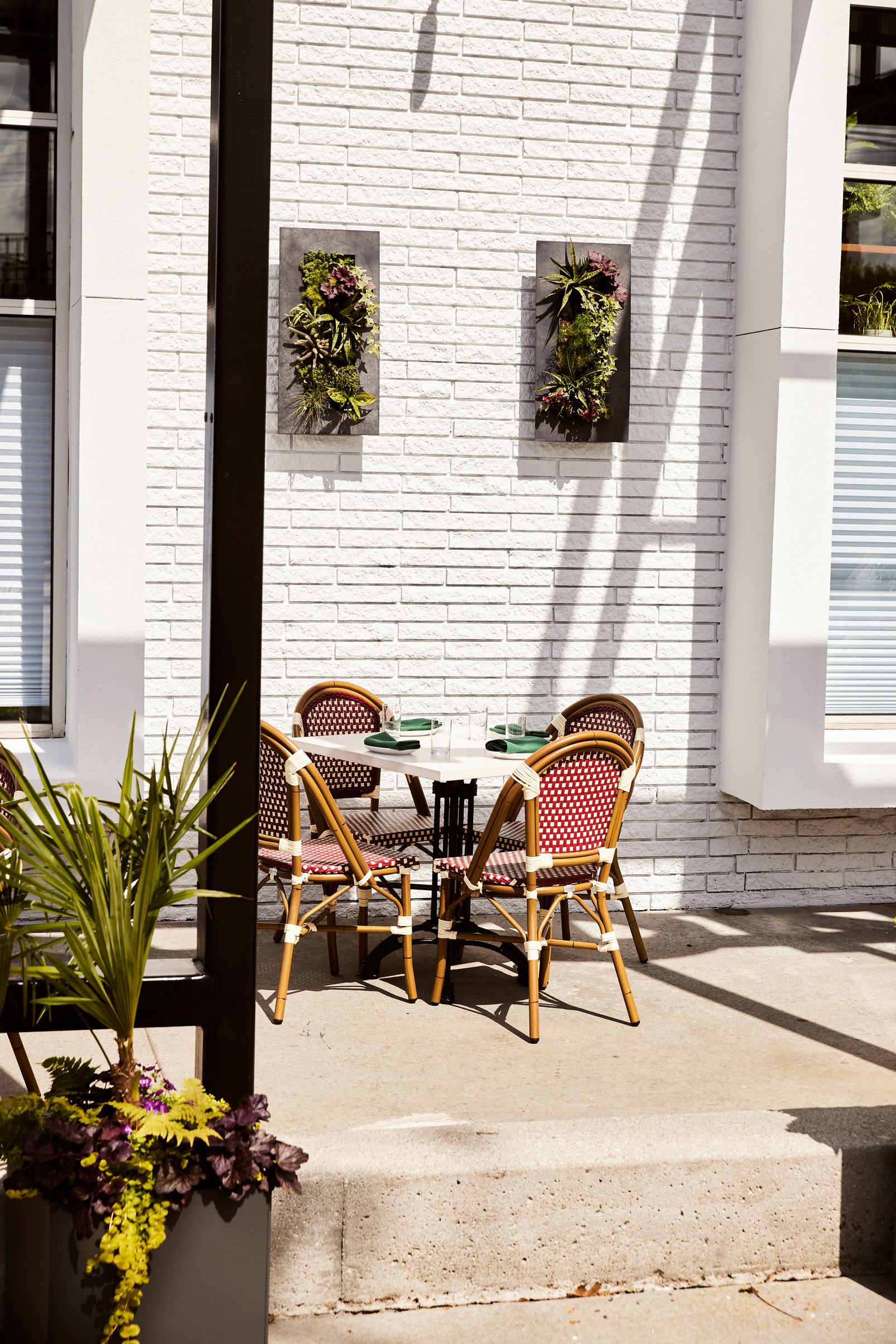 A photo of a charming outdoor dining area with a white brick wall. Two vertical planters with lush greenery hang on the wall. A round table with four red and white striped chairs is set for a meal. Sunlight casts dappled shadows on the patio, creating a warm and inviting atmosphere.