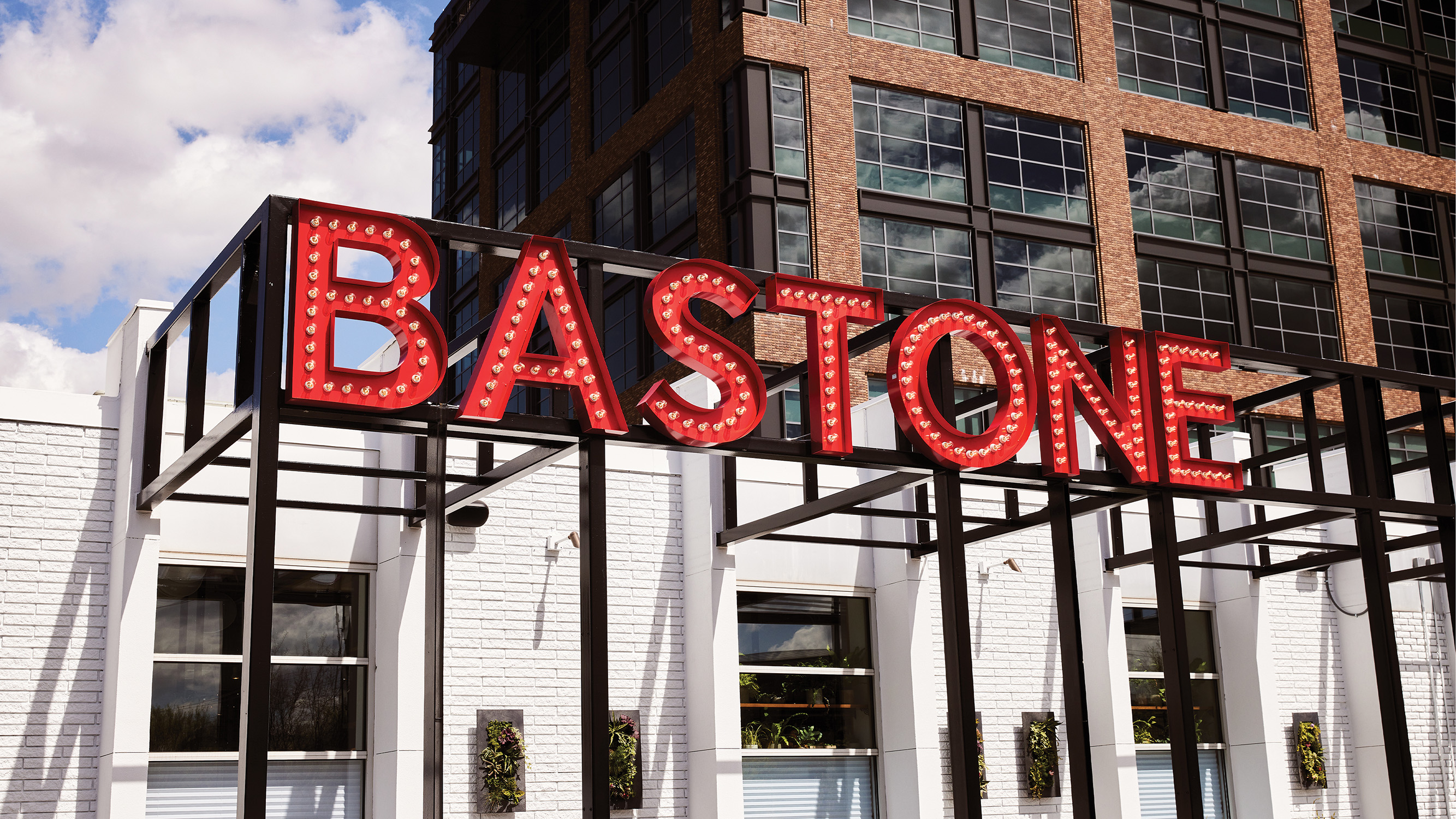 A photo of the Bastone restaurant storefront. The restaurant name is displayed in large, illuminated red letters on a metal frame structure above the entrance. The building has a modern design with large windows and a brick facade.