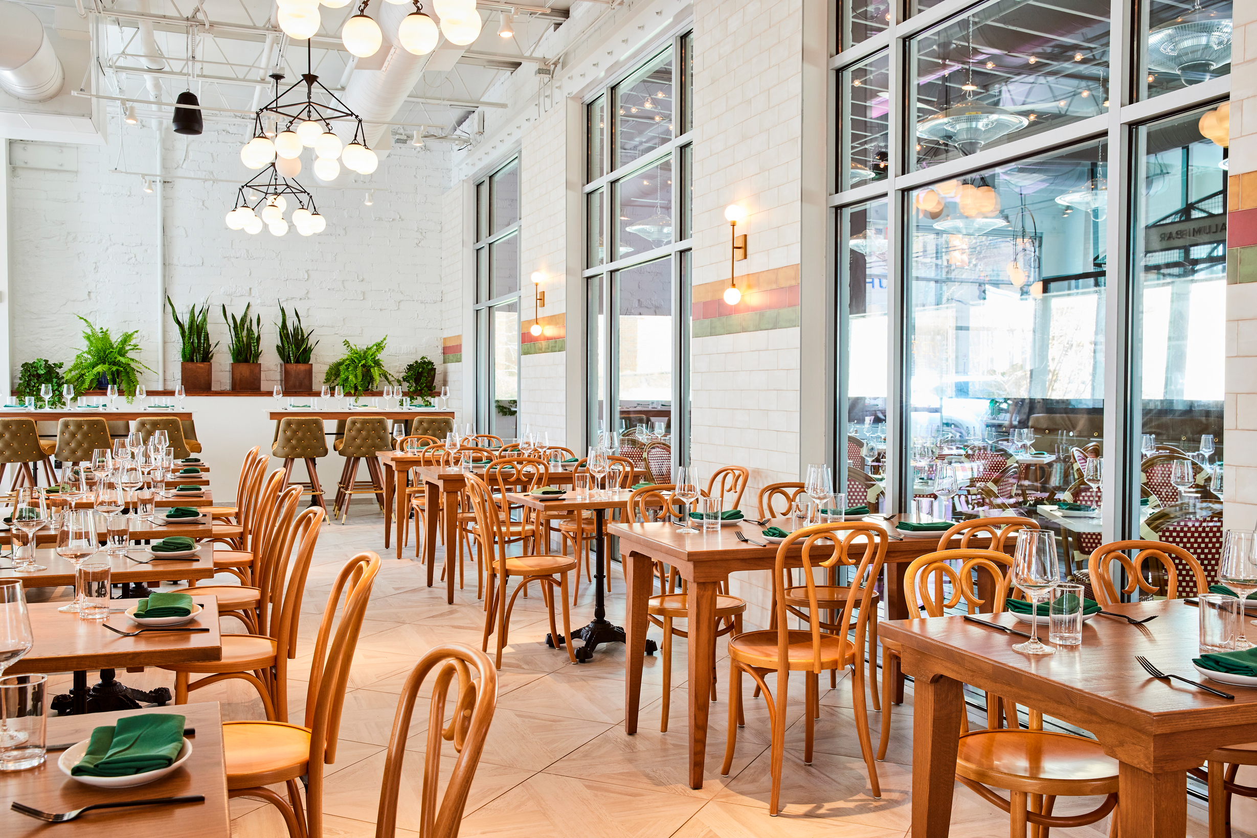 A photo of a modern and inviting Bastone interior. The space features a mix of wooden tables and chairs, with large windows that bring in plenty of natural light. The walls are white brick, and there are hanging plants and decorative lighting fixtures adding warmth and character.