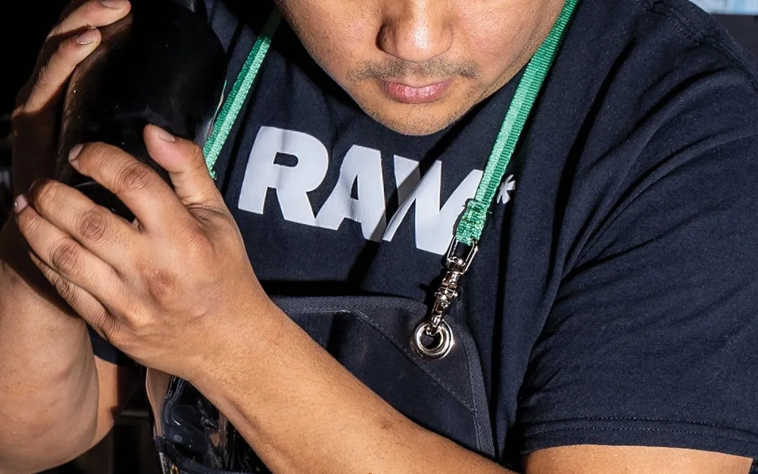 Bartender wearing a black apron and green strap around neck, holding a cocktail shaker, wearing a black shirt with the word "RAW" partially visible.