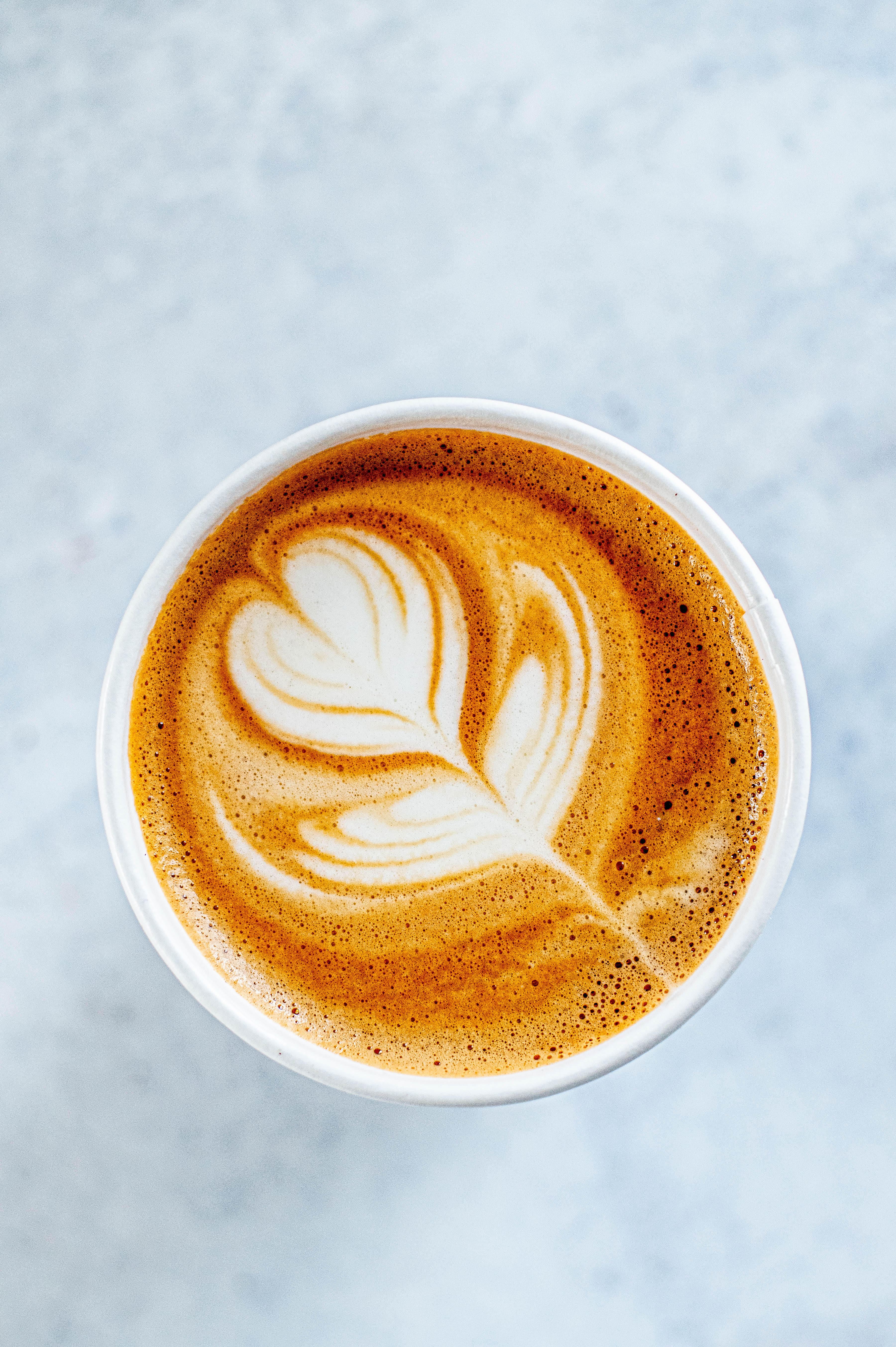 Close-up of a white cup containing a latte with a delicate, heart-shaped design created in the foam, set against a light marble background.