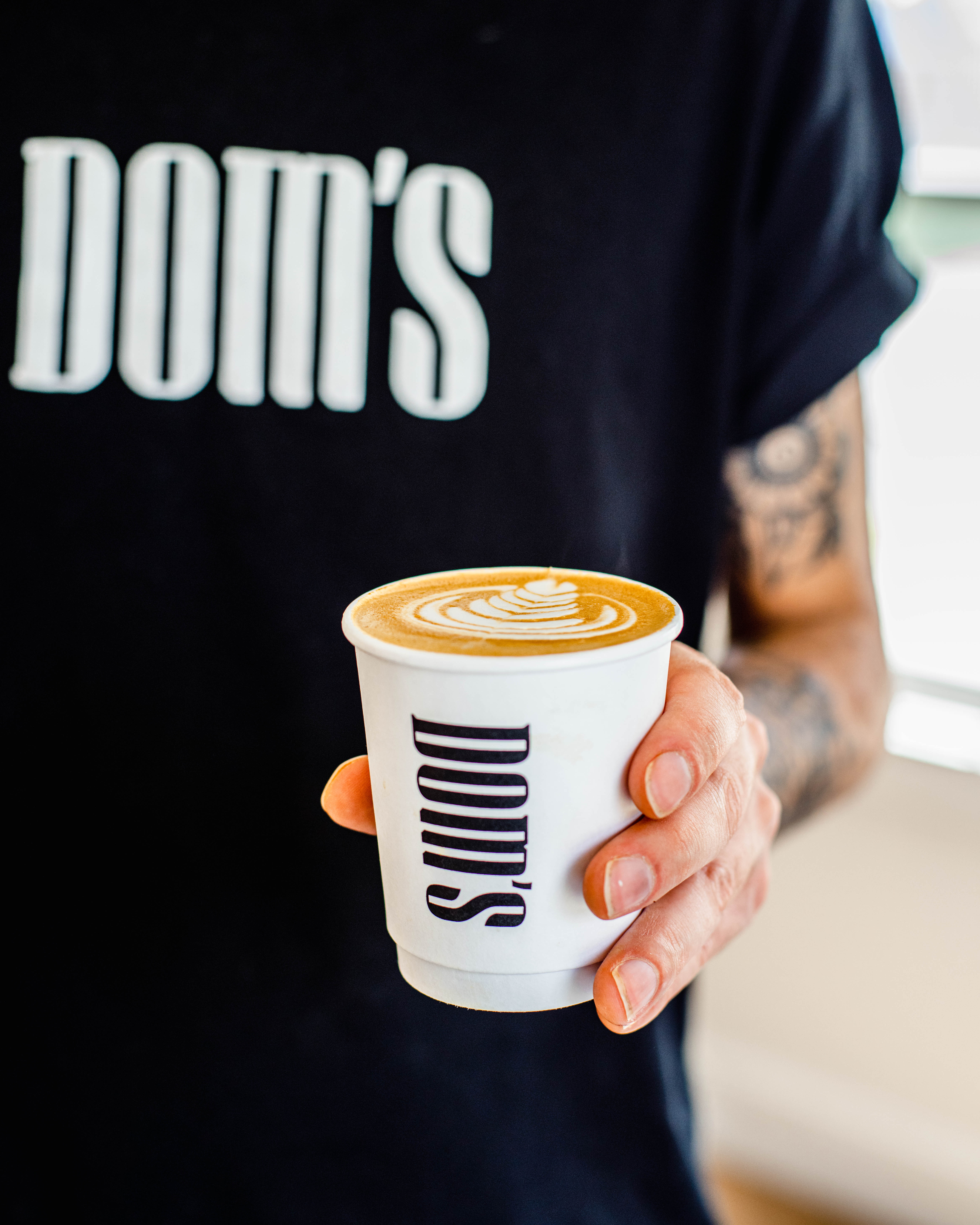 Close-up of a hand holding a Dom's coffee cup, with the "Dom's" logo prominently displayed, against a background of a person wearing a Dom's branded t-shirt.