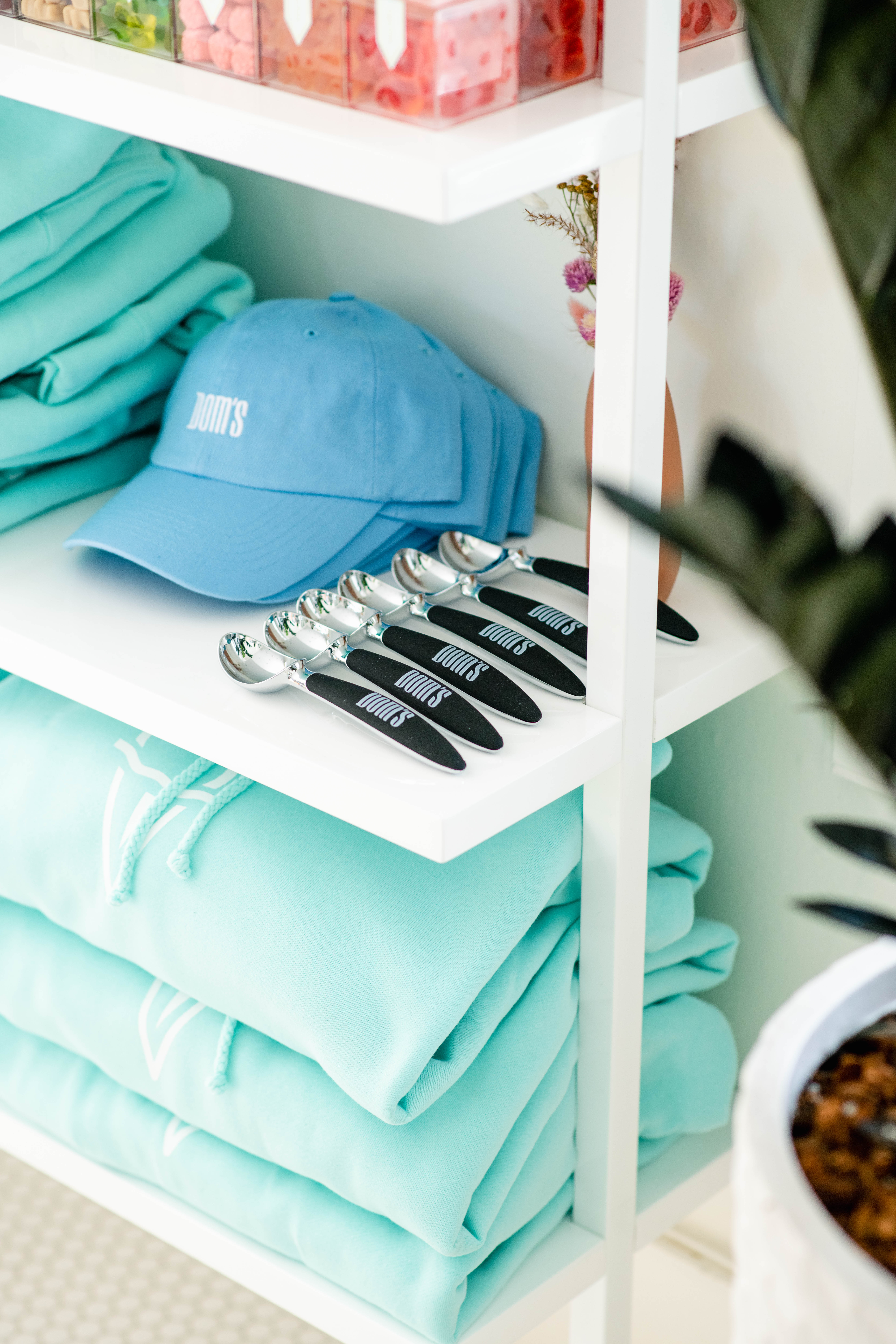 Close-up of a white retail shelf displaying folded light teal apparel with the "Dom's" logo, a matching baseball cap, and a row of branded spoons, showcasing the brand's merchandise and visual identity.