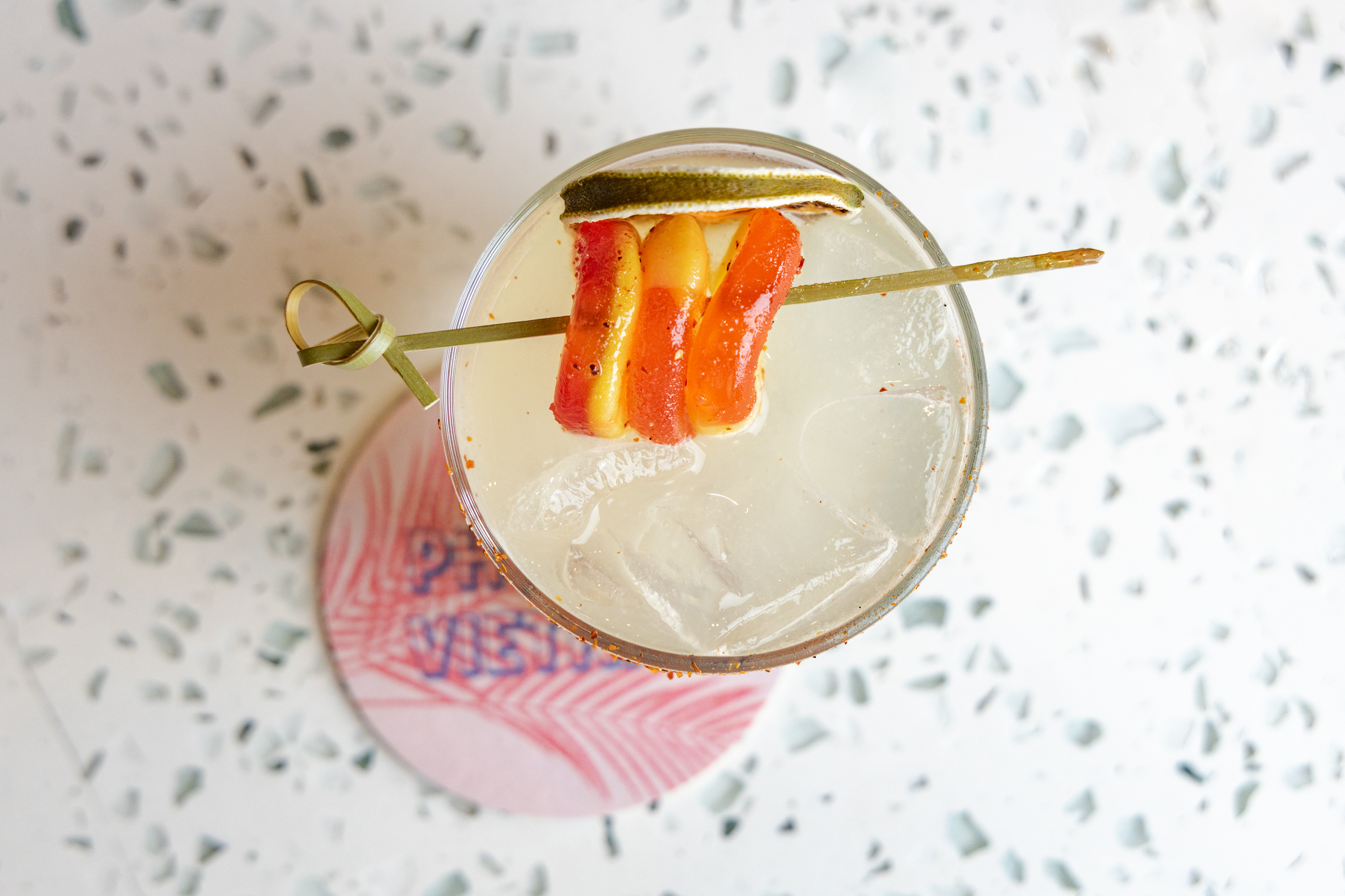 Flat lay photograph of a margarita cocktail in a salt-rimmed glass, garnished with red chili peppers and a lime wheel, resting on a branded coaster with a geometric pattern.