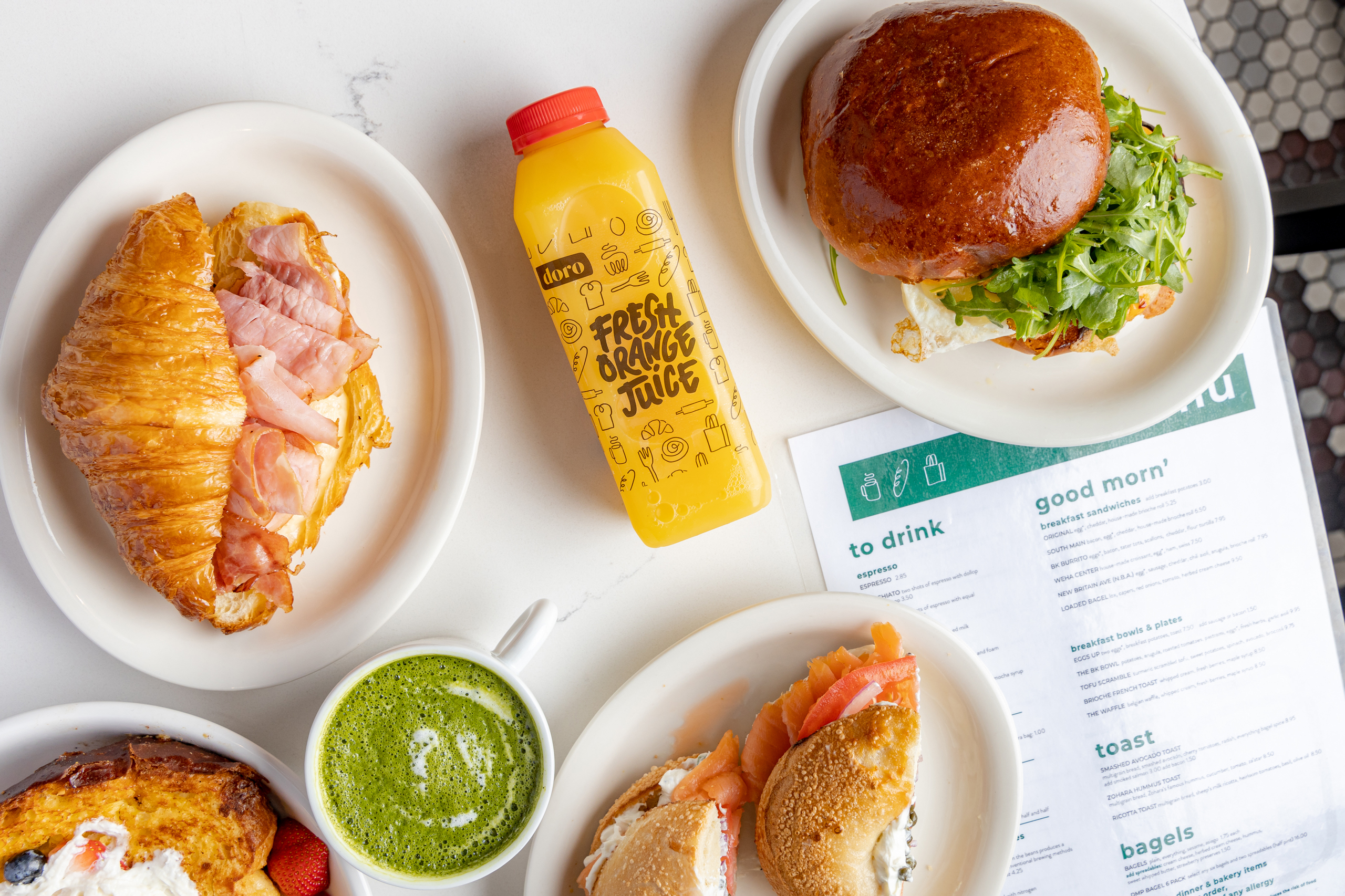 A photo showcasing a vibrant breakfast spread at Doro Marketplace. The table features pastries, sandwiches, juice, and tea, with the Doro Marketplace menu on display.