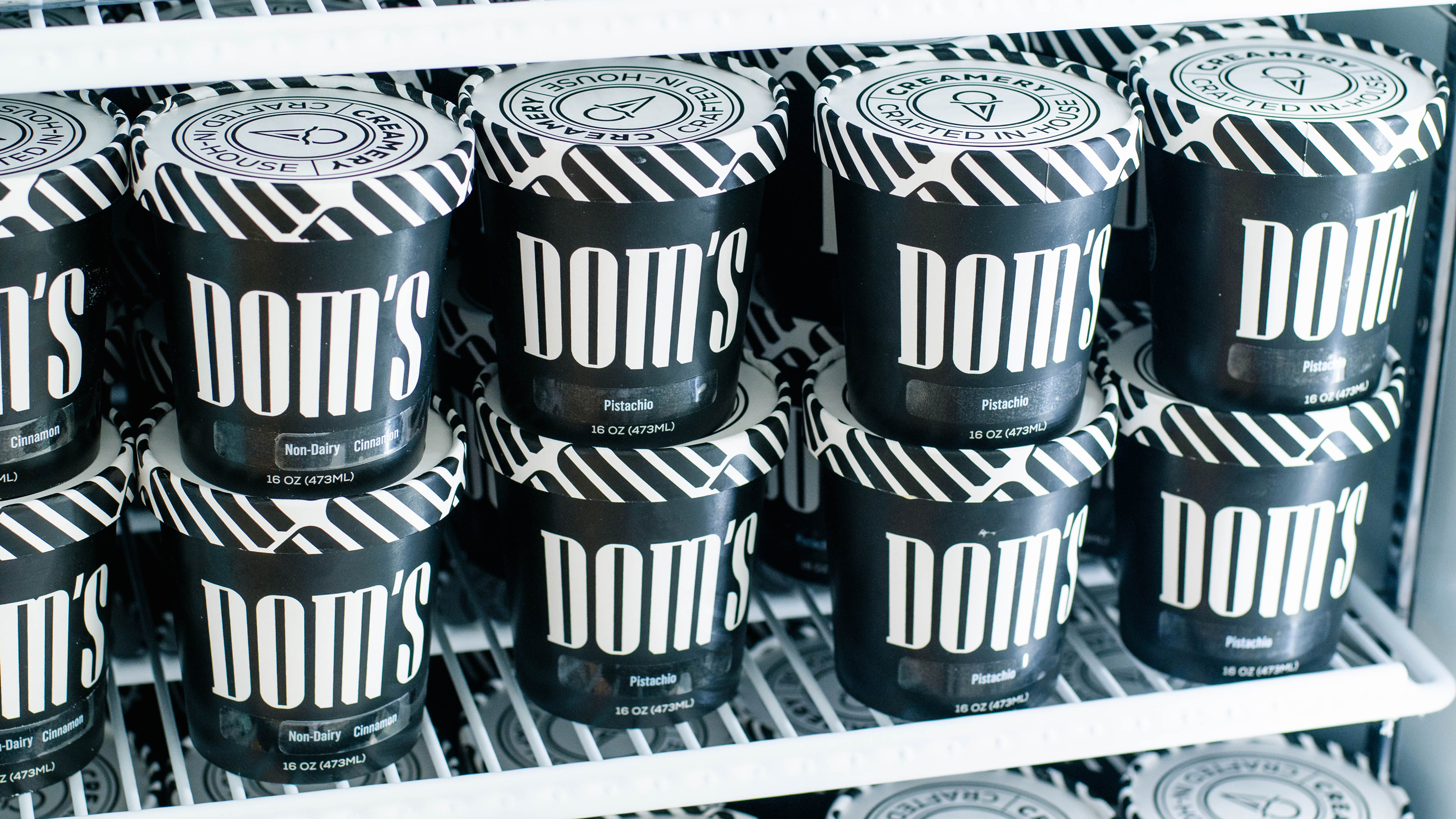 Close-up of several black ice cream containers stacked in a refrigerator display, featuring the "Dom's" logo in a bold white font and flavor labels, highlighting the product's branding and presentation.