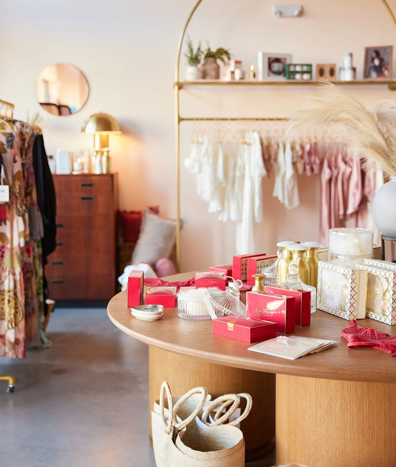 Interior view of a curated retail space featuring a round display table with boxed items and decorative objects, a clothing rack showcasing garments in soft hues, and a warm, inviting ambiance created by the lighting and color palette.