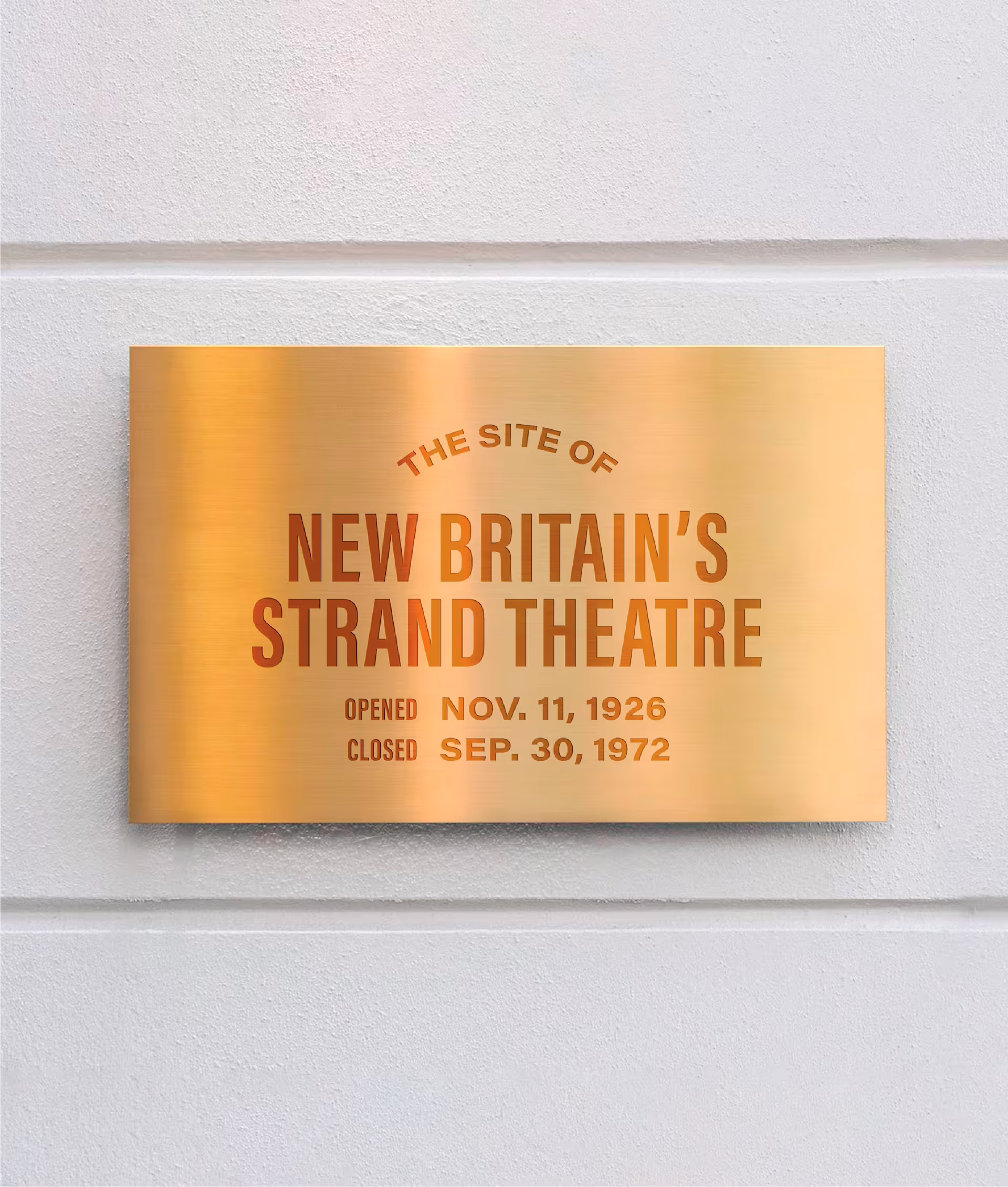 Rectangular bronze plaque mounted on a textured wall, commemorating the location of the former Strand Theatre with engraved text detailing its opening and closing dates.