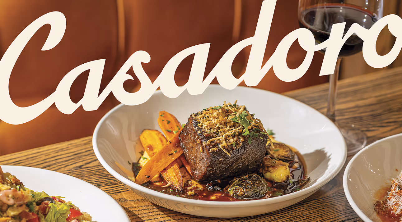 A stylized product shot featuring two ruby-red cocktails on a wooden board, garnished with lime wedges and fresh mint. The Casadoro logo is overlaid in a flowing script typeface, creating a sense of elegance and sophistication. The composition is complemented by strategically placed citrus fruit, adding pops of color and texture.