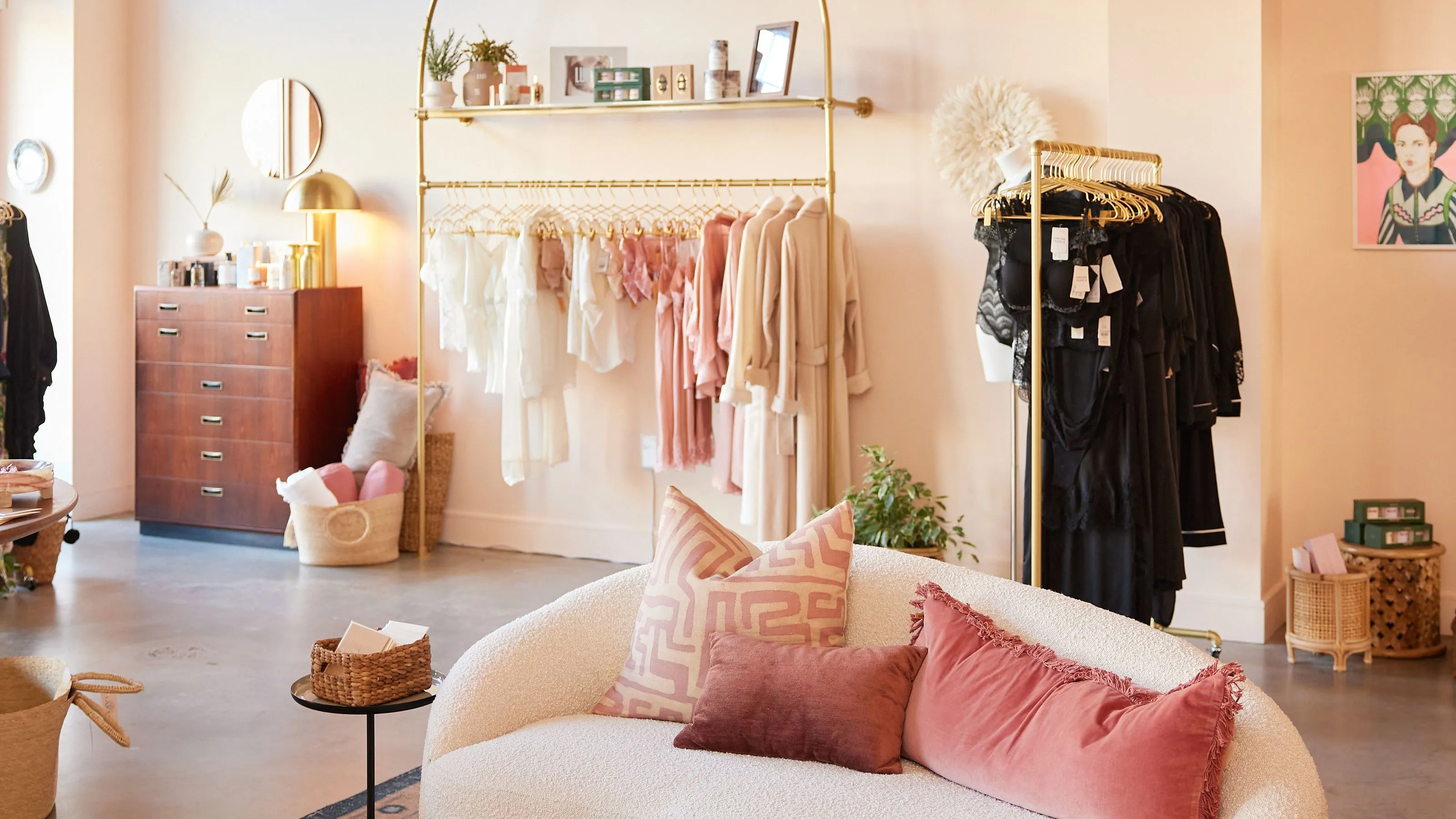 LaSource's interior featuring a curved, cream-colored sofa with decorative pillows, a clothing rack displaying garments in neutral tones, and a shelving unit with decorative items, all contributing to a curated and inviting shopping environment.