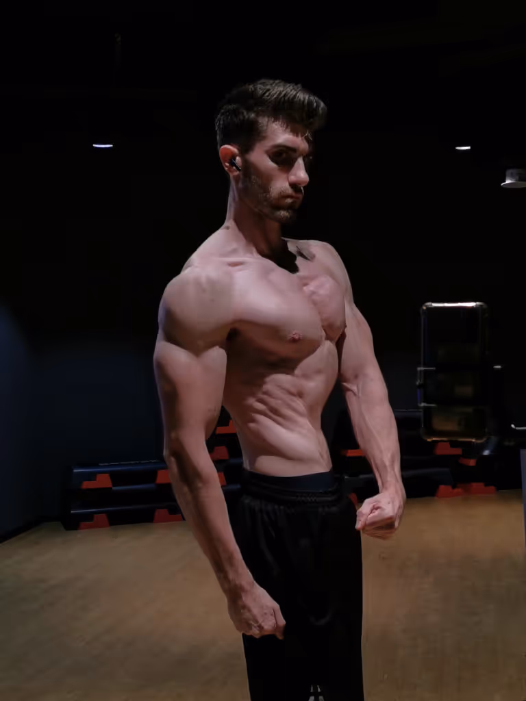 Shirtless muscular man posing in a dimly lit gym with black and red workout equipment in the background.