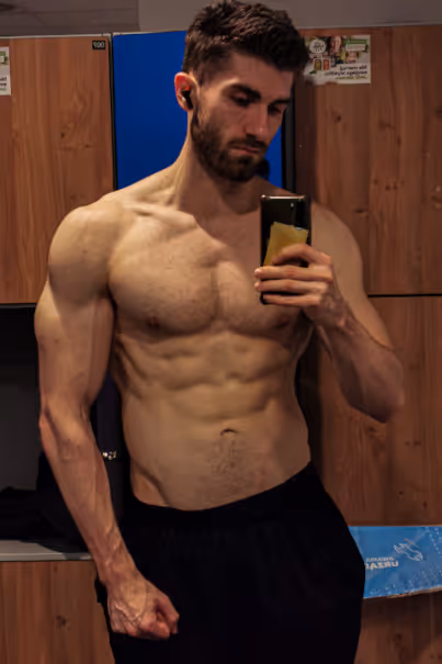 Muscular man with earbuds taking a shirtless mirror selfie in a locker room.