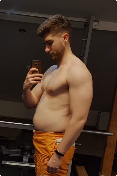 Shirtless young man taking a selfie in a gym mirror, wearing orange shorts and a fitness tracker.