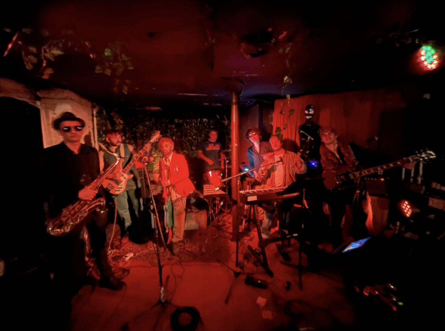 Seven-member band performing in a dimly lit venue with red lighting, featuring instruments like saxophone, bass guitar, drums, keyboard, and electric guitar.