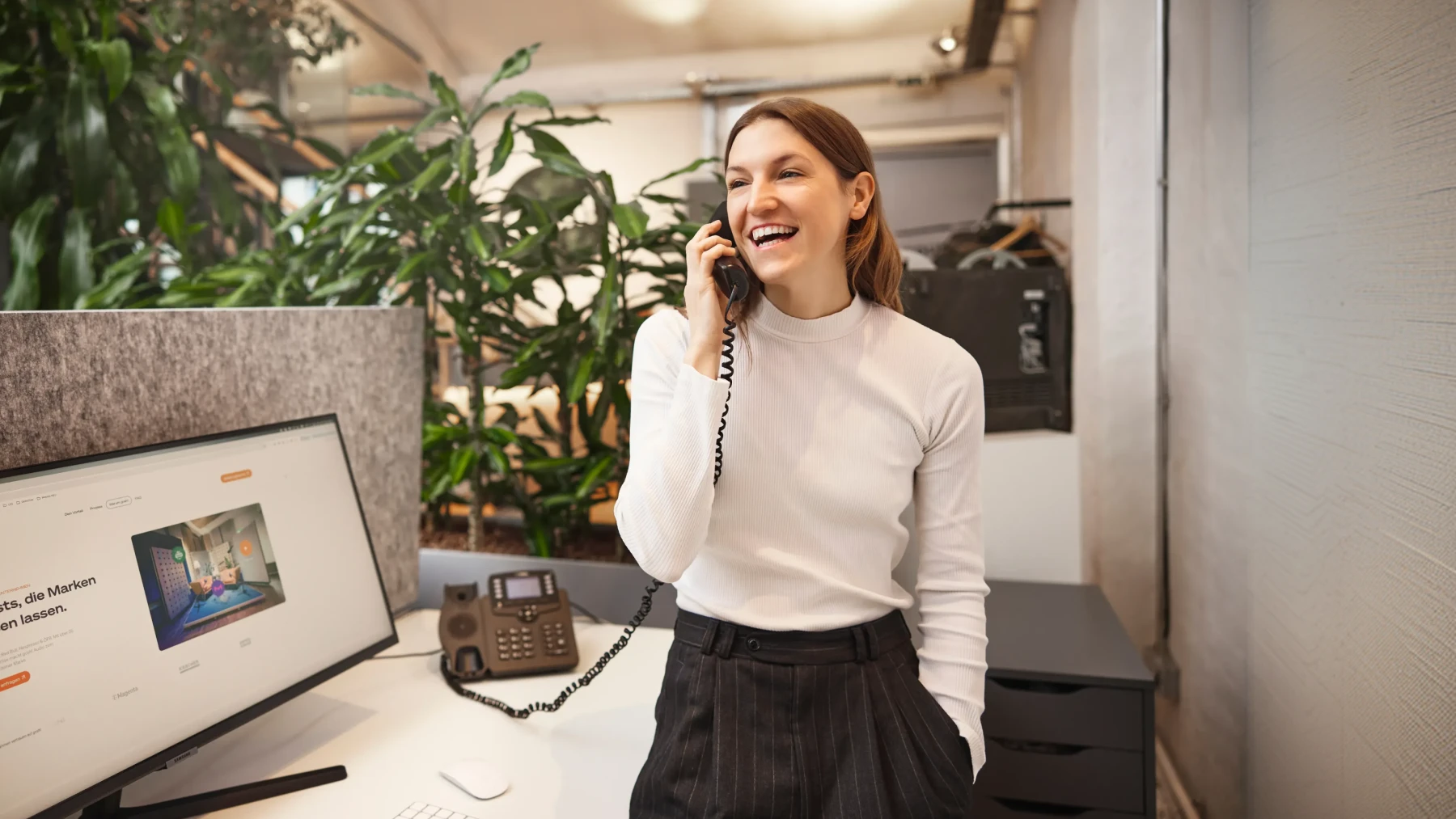 Lächelnde Frau am Arbeitsplatz telefoniert mit Festnetz-Telefon vor Computerbildschirm und Pflanzen.