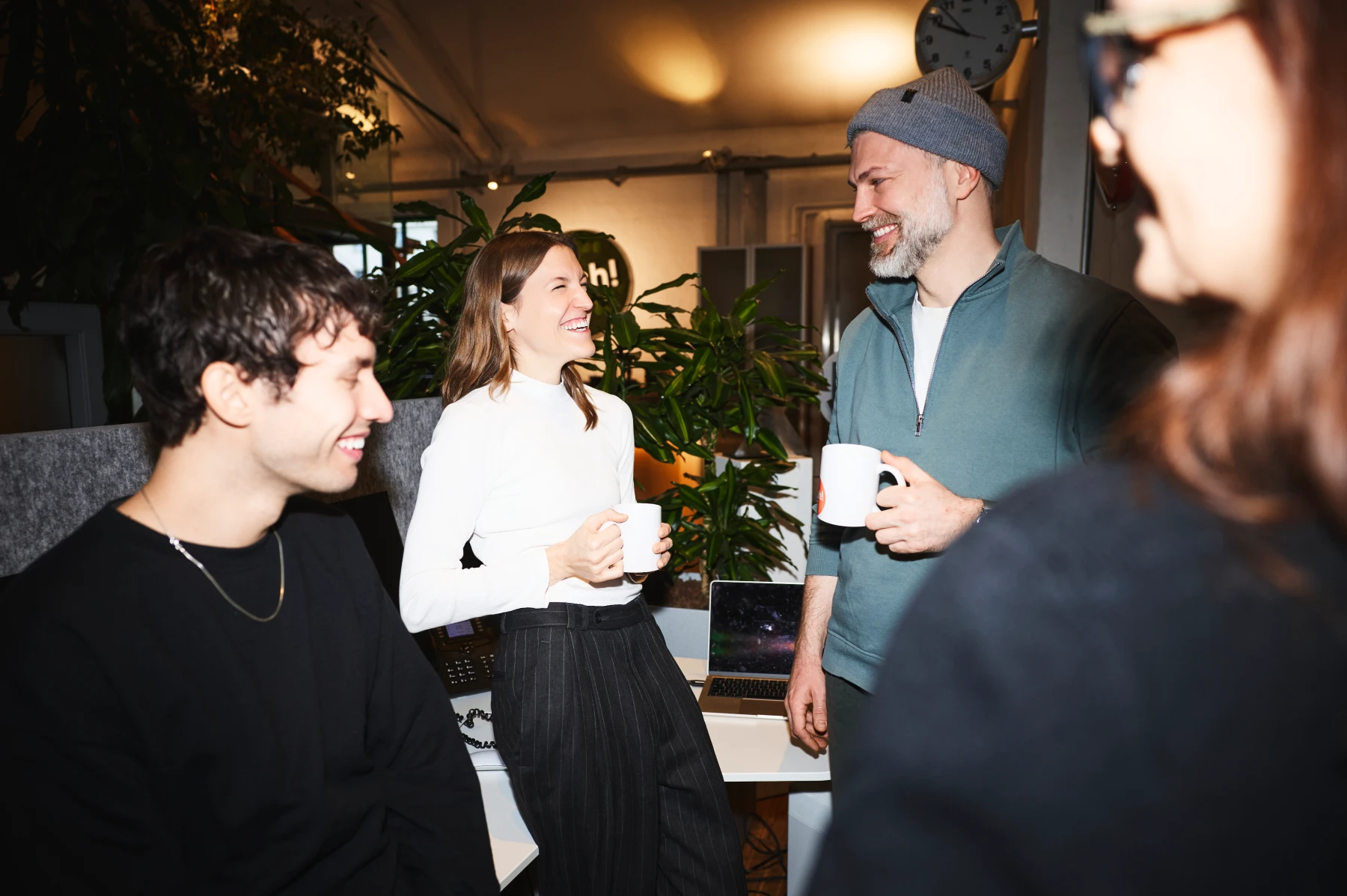 Vier Personen lachen und trinken Kaffee in einem modernen Büro vor einem Laptop und Pflanzen.