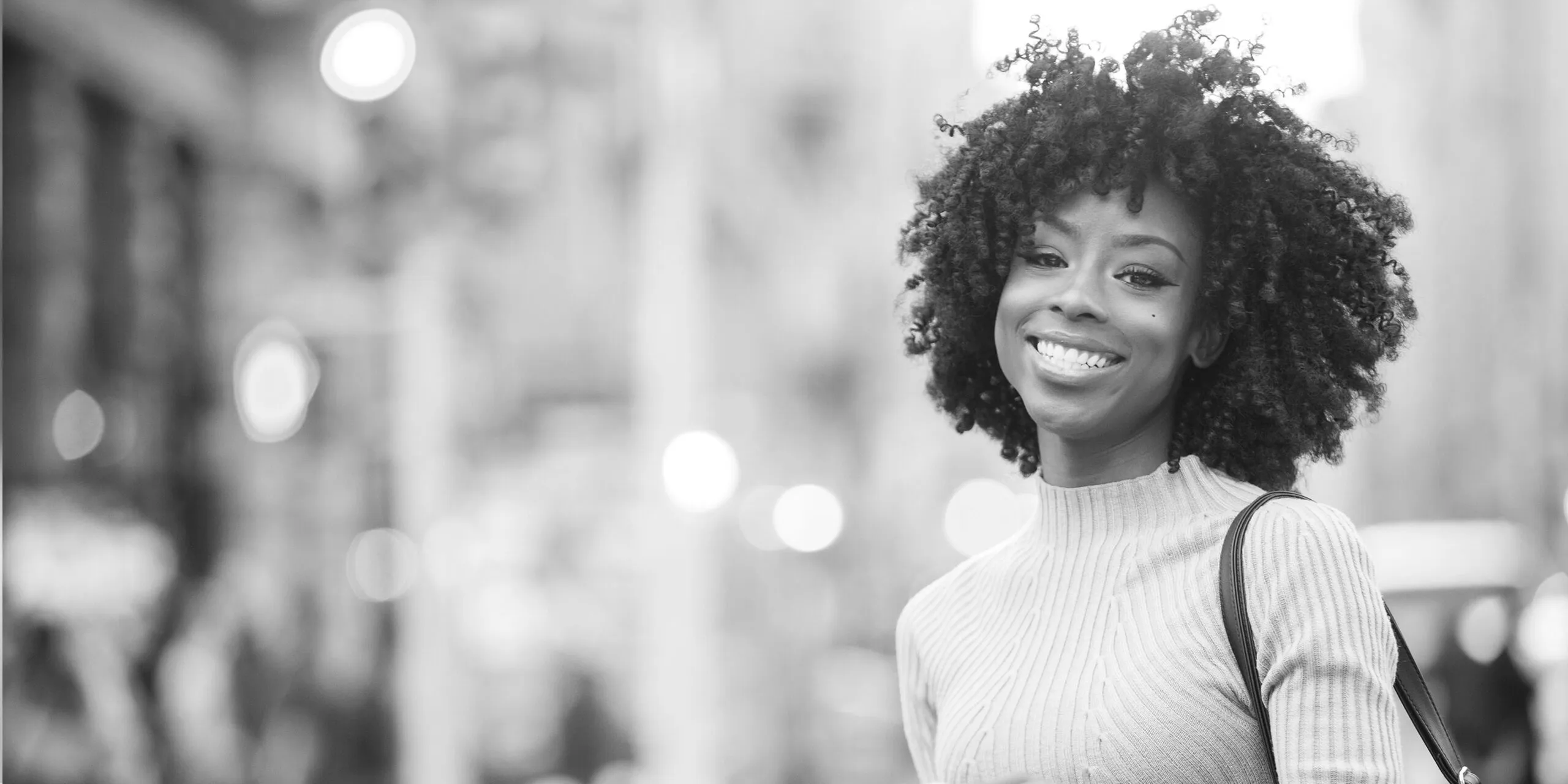Smiling woman with curly hair wearing a ribbed sweater and crossbody bag on a city street.