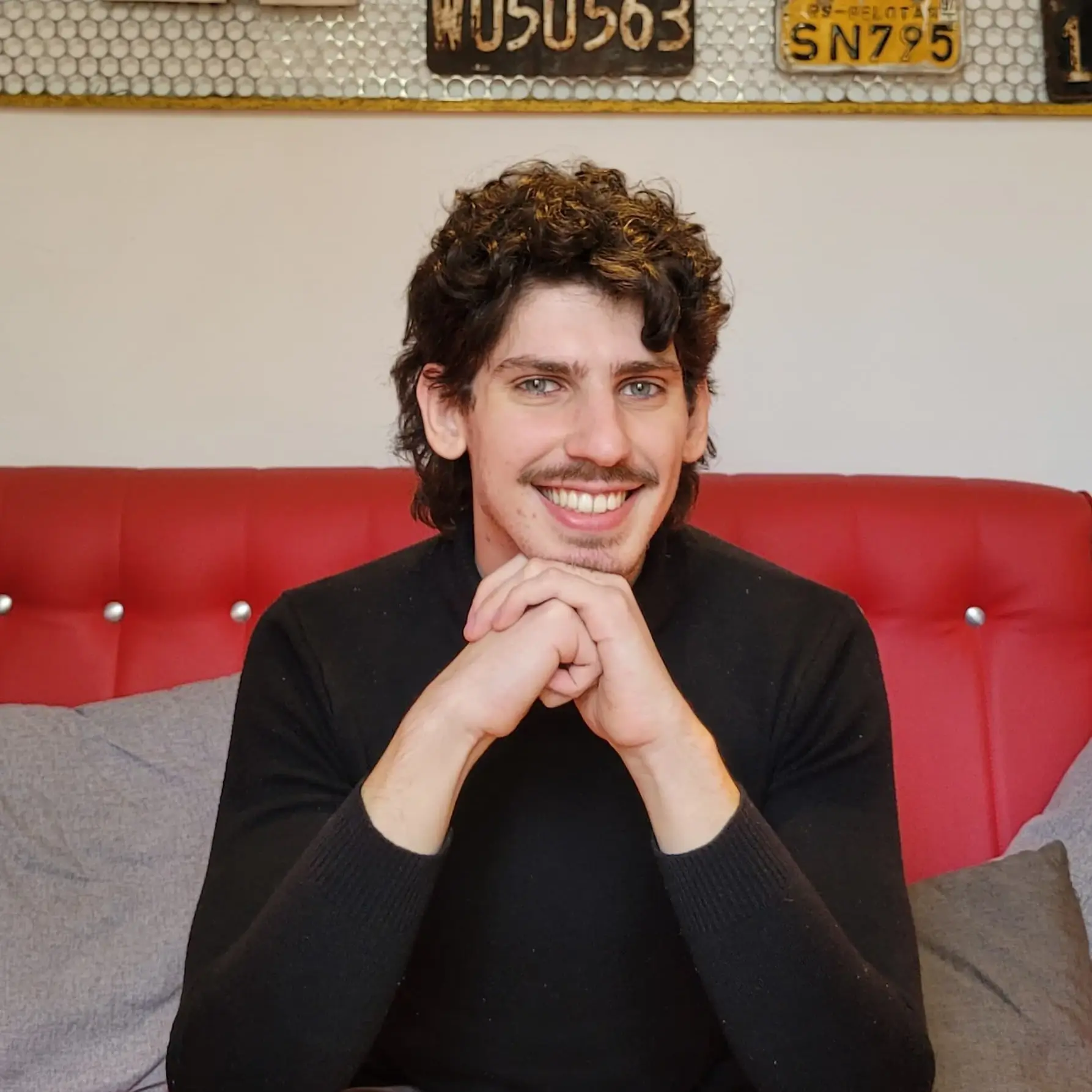 Imagem do proprietário da Rezer Estúdio, Arthur Rezer, sentado, com uma blusa preta de gola alta, sorrindo para a câmera, com o queixo apoiado nas mãos, de cabelo cacheado e olhos azuis.
O fundo tem um quadro de leitura ilegível apoiado na parede logo atrás do homem, que está sentado em um sofá vermelho.