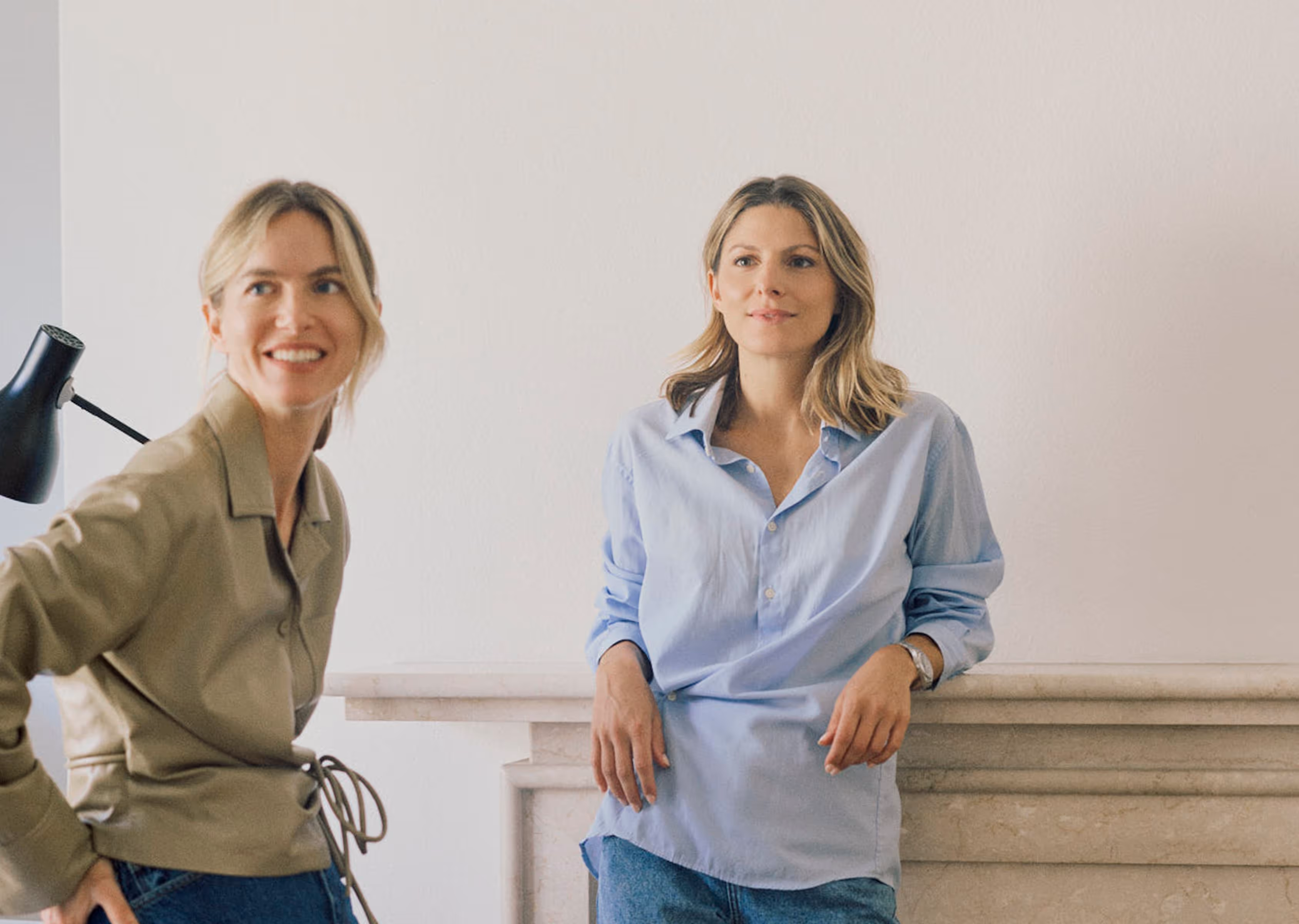 Zwei Frauen in legerer Kleidung lehnen entspannt an einer hellen Wand und einem Kamin.