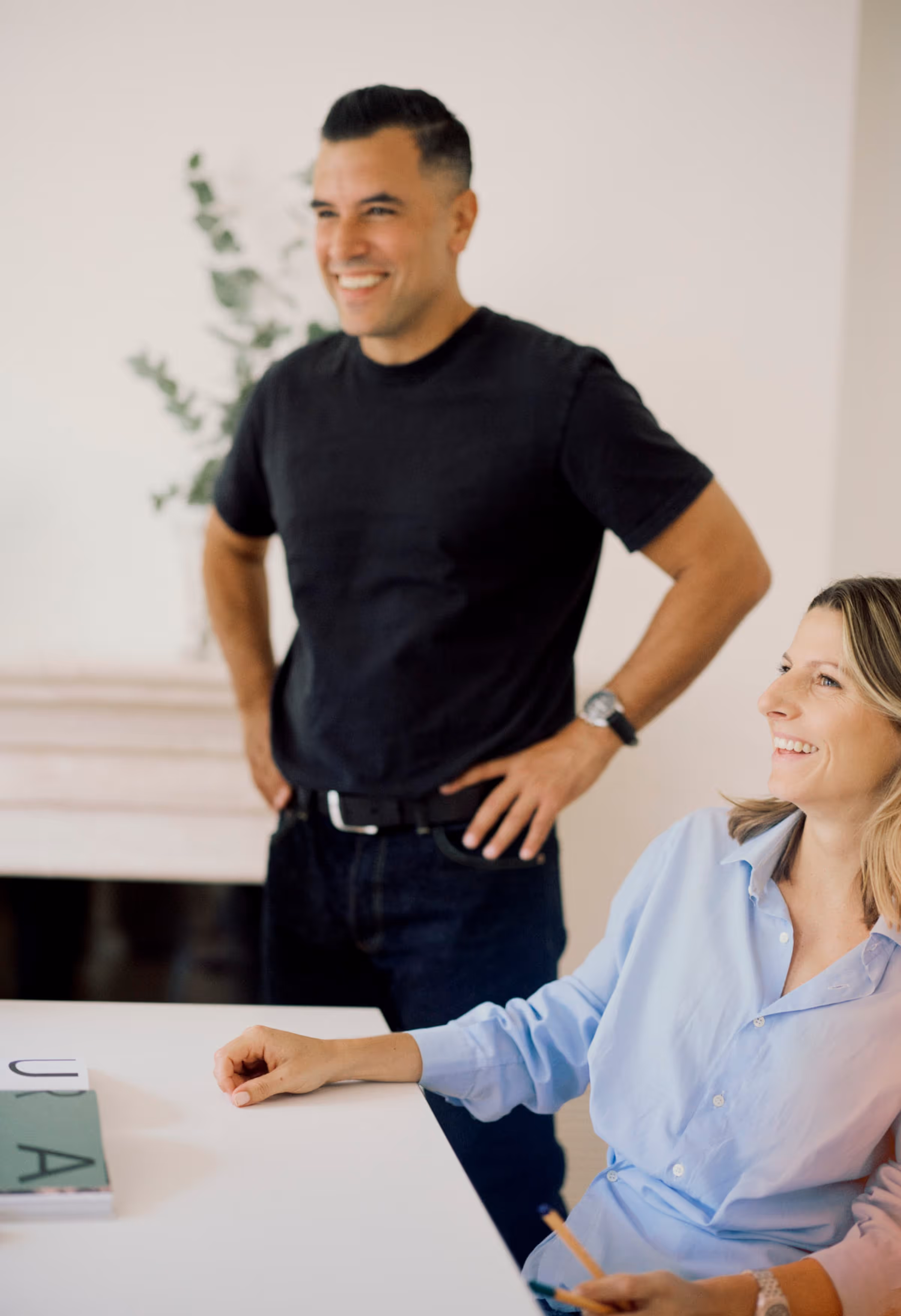Ein lächelnder Mann in schwarzem T-Shirt steht mit Händen in der Hüfte, eine lächelnde Frau in blauer Bluse sitzt am Tisch mit Stift in der Hand.