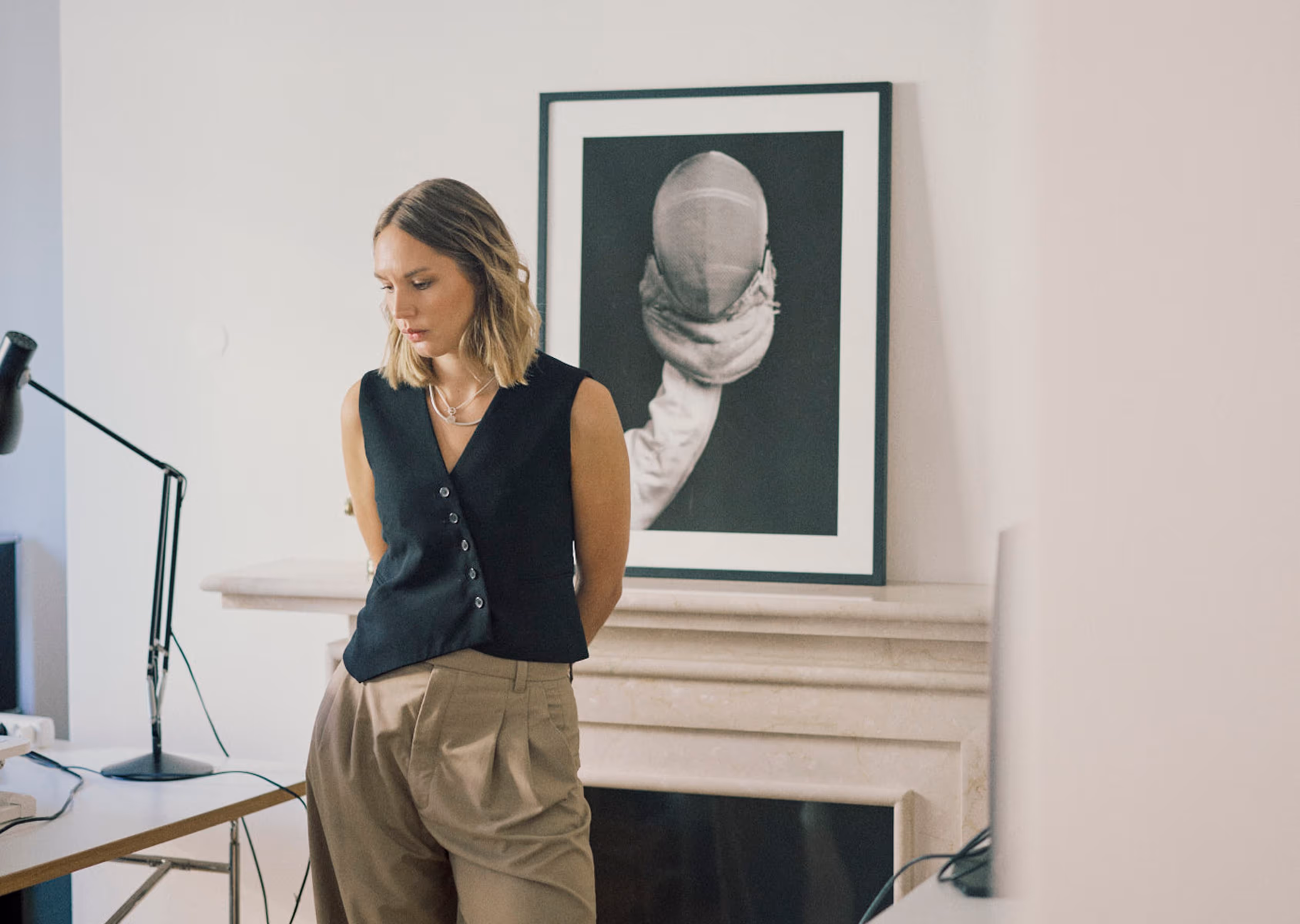 Frau in schwarzer Weste und beiger Hose lehnt vor einem Kamin mit einem gerahmten Schwarzweißfoto von einer Person mit Fechtausrüstung.