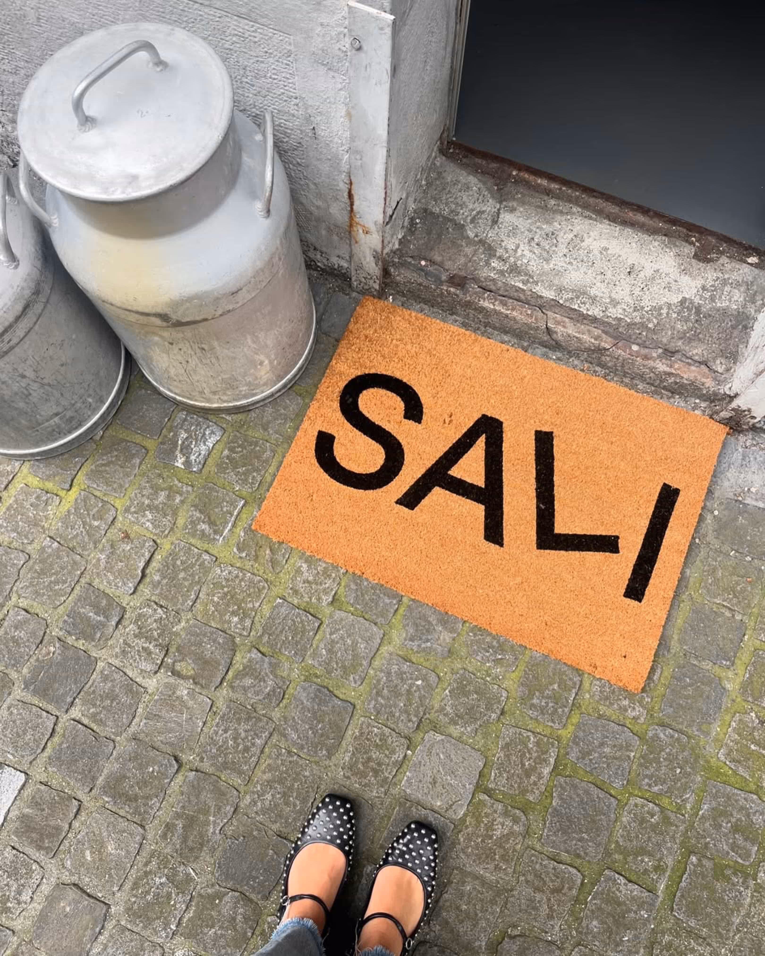 Fussteppich mit der Aufschrift 'SALI' vor einer Tür, daneben zwei silberne Milchkanister auf Kopfsteinpflaster, mit Blick auf Schuhe mit schwarzen Nieten.