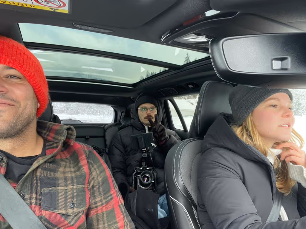 Drei Personen in einem Auto mit Schiebedach, der Mann vorne links trägt eine rote Mütze, die Frau vorne rechts eine graue Mütze, und die Person hinten hält eine Kamera.