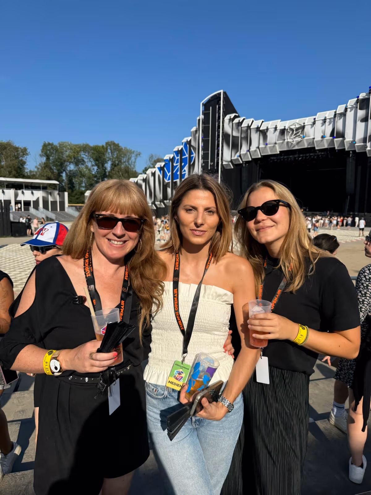 Drei Frauen mit Sonnenbrillen und Festival-Ausweisen posieren vor einer Bühnenkulisse bei klar blauem Himmel.