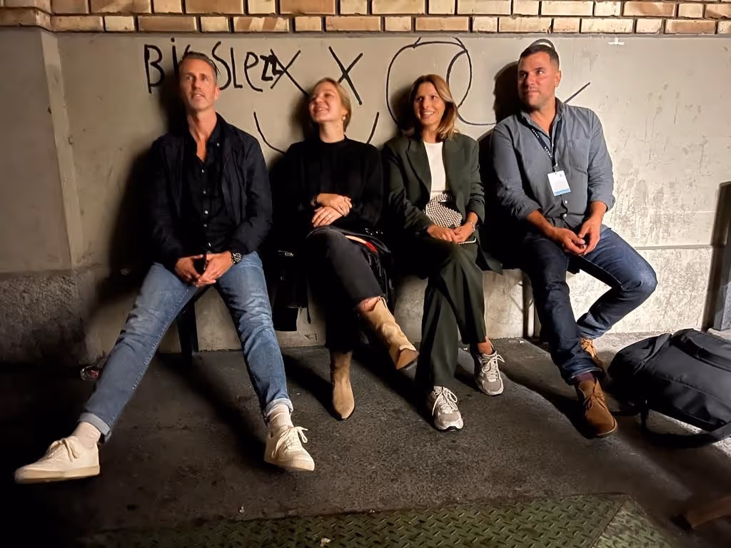 Vier Personen sitzen an einer Wand mit Beton und Backstein, lächelnd und entspannt, mit Schreibschrift-Graffiti an der Wand.